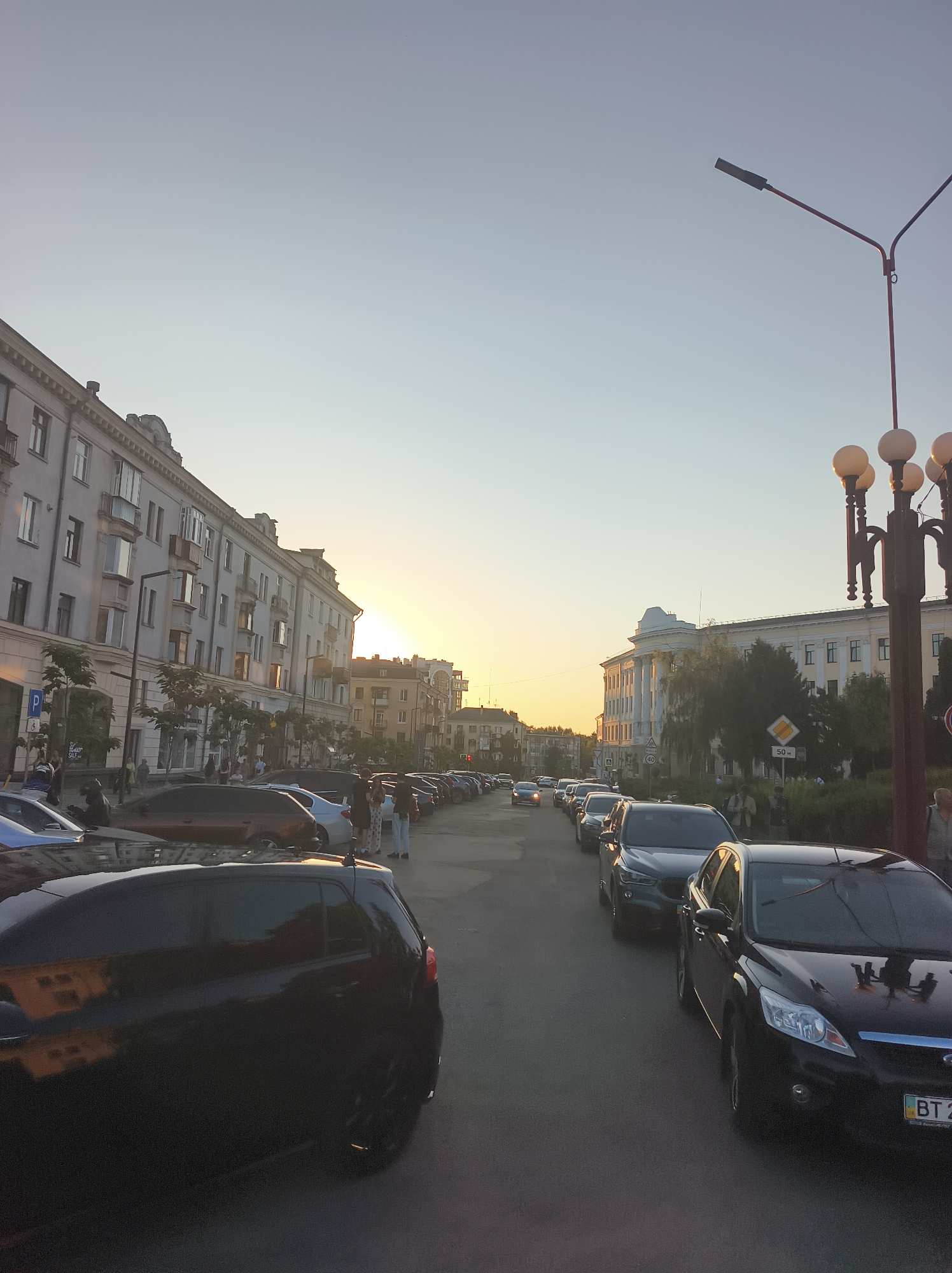 This media file captures a street scene in Ternopil, Ukraine, during what appears to be the golden hour as the sun sets. The sky is a clear, pale blue, transitioning to a warm yellow hue near the horizon where the sun is partially obscured by buildings. The scene is dominated by a street lined with parked cars and a few pedestrians.
On the left side of the street, a long, multi-story building with a classical facade stands, featuring numerous windows and balconies. Trees with sparse foliage are planted along the sidewalk. The right side of the street features a large, white building with columns, suggesting it might be a public institution or historical landmark. A street lamp with multiple globes stands prominently in the foreground on the right, illuminated against the twilight sky.
Several cars are visible, parked closely together on both sides of the street and in the middle lane, indicating a busy urban environment. Some people are walking along the sidewalks, while others are standing near the parked cars, engaged in conversation. The overall atmosphere appears calm and relaxed, typical of a late afternoon or early evening in a city.
Visible text includes a parking sign on the left and a speed limit sign of "50 km/h" on the right. A license plate on a car in the foreground on the right shows "BT 2". The light conditions suggest it is late afternoon or early evening.
