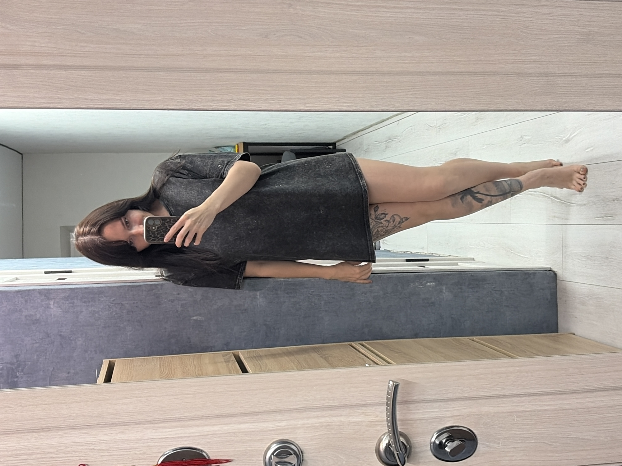 The image displays a full-body mirror selfie of a person, likely female, captured indoors. The subject is reflected sideways in what appears to be a full-length mirror, positioned as if standing with their head towards the upper left of the frame. They have long, dark hair and are wearing an oversized dark grey or black t-shirt dress. Their bare legs are visible, adorned with tattoos, including a prominent leafy or floral design on the upper left thigh and another, less defined, tattoo on the lower right leg.
The person is holding a smartphone with a distinctive leopard print case, aiming it towards the mirror to take the photograph. Their eyes are directed towards the phone screen, indicating focus on the selfie process.
The setting reveals elements of an indoor environment. Below the mirror, there's a light-colored wooden surface, resembling a door or cabinet, fitted with a silver-toned metal handle and two circular lock mechanisms. In the reflection, behind the person, a grey-blue surface is visible, possibly a wall or another piece of furniture, along with a light-colored wooden floor. The lighting is consistent with an indoor setting, and no specific time of day can be determined. There is no visible text in the image. The location context for this media file is Chaikovsky, Russia.