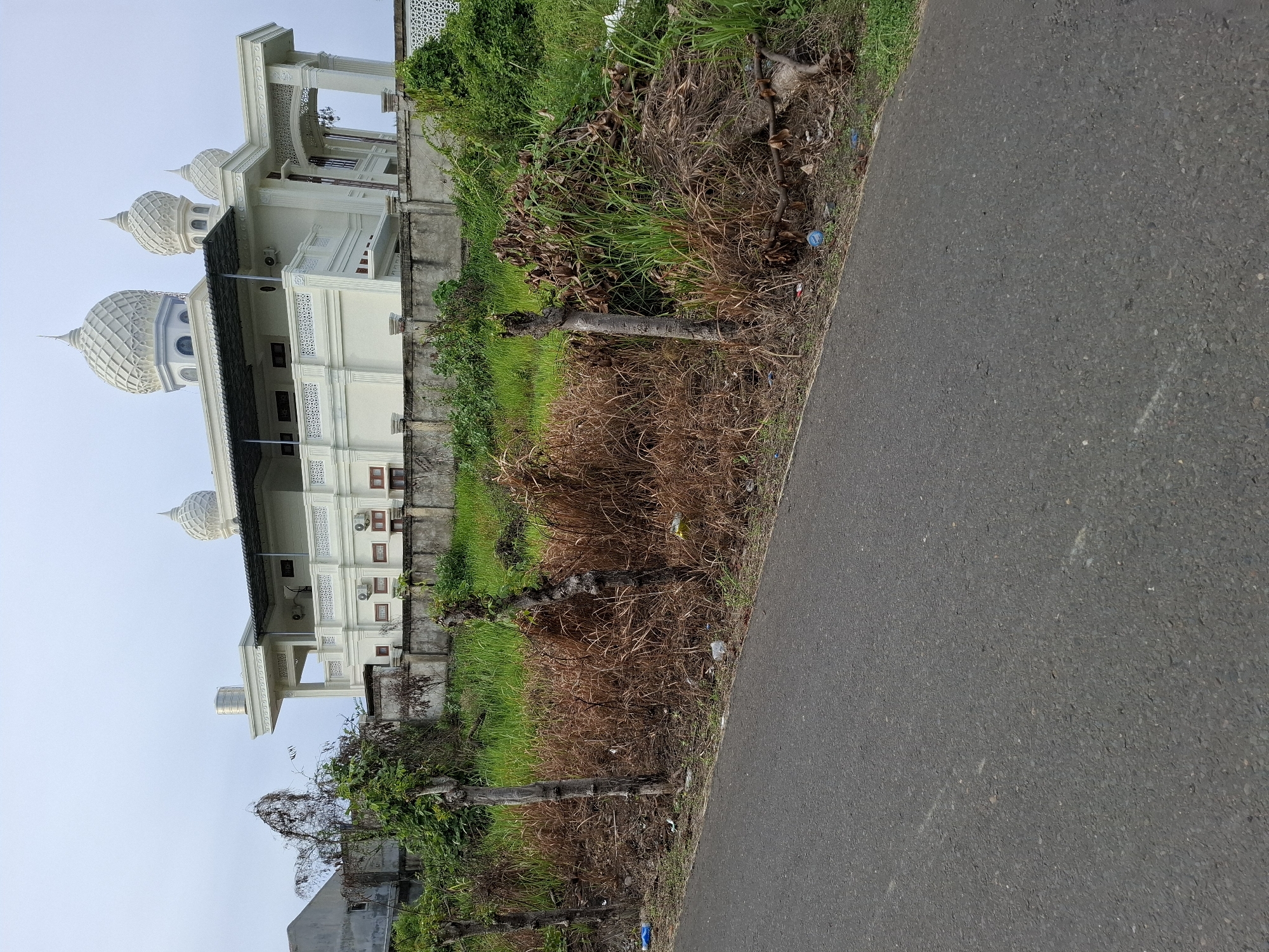 The image shows a view of Masjid Islamic Center Lhokseumawe, a mosque located in Lhokseumawe, Indonesia. The mosque is a white building with multiple onion-shaped domes. The side of the mosque is visible behind a low wall, with grass and vegetation growing between the wall and the road. An asphalt road stretches across the foreground of the image. The sky is overcast.