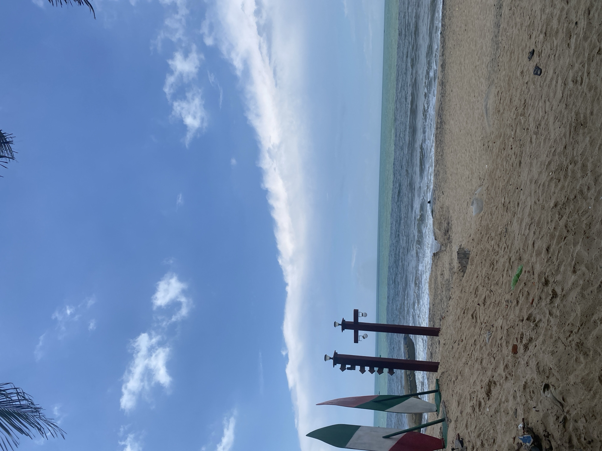 This is an eye-level, medium shot of a tropical beach scene, oriented vertically. The left side of the image is dominated by a bright blue sky with scattered white clouds, some of which form a long, thin band stretching across the frame. In the upper left corner, the fronds of a palm tree are visible.
The right side of the image depicts a sandy beach leading to the ocean. The sand is a light brown and appears somewhat damp near the water. Gentle waves with white foam are rolling onto the shore. In the distance, the water is a muted green-blue, and the horizon is a hazy line where the sea meets the sky.
In the foreground, two wooden posts stand on the beach, each with what appear to be decorative lights or speakers attached to the top. Between these posts, and partially visible at the bottom of the frame, are two colorful objects that resemble surfboards or decorative panels, painted with red, white, and green sections.
Scattered on the sand are a few pieces of debris, including what looks like a green plastic bag and a pair of dark flip-flops. There are no people visible in the frame. The overall impression is one of a tranquil beach setting on a day with partly cloudy skies, likely during daylight hours. The location cues, such as the palm tree and the surfboards, suggest a warm, possibly humid climate.