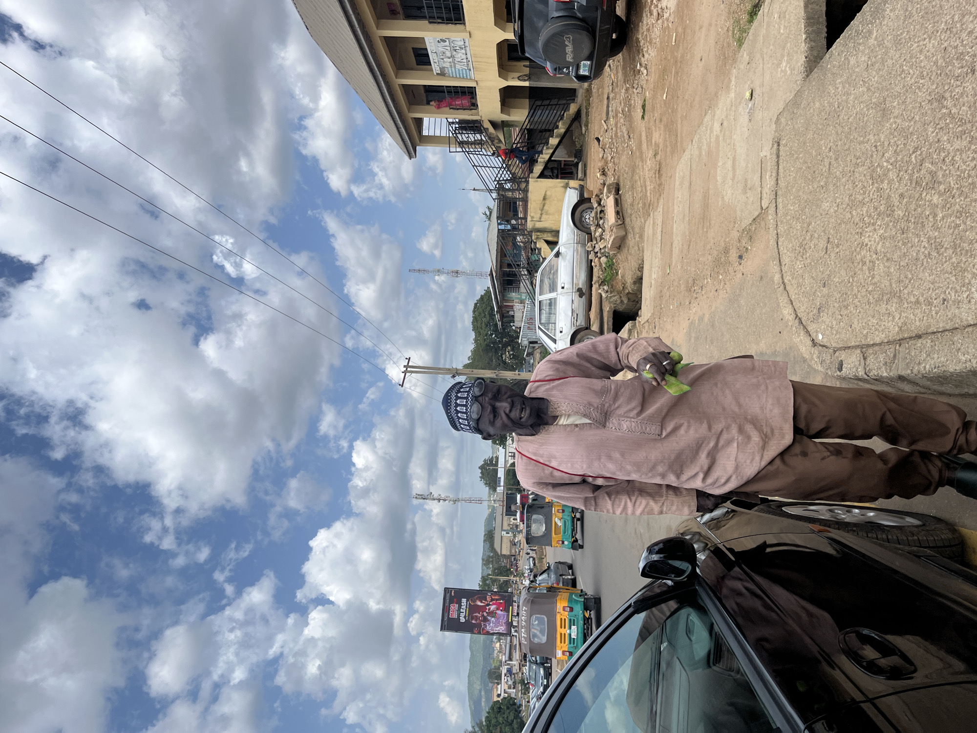 The photograph depicts a street scene in Rafin-Par, Nigeria. In the foreground, an elderly man wearing traditional attire, including a pink tunic and brown trousers, stands on the sidewalk. He has a dark skin tone, is bald, and wears a dark cap and glasses. He is holding a small green object in his right hand. To his left, a black car is parked.
The background reveals a busy street with several auto-rickshaws (also known as Keke-NAPEPs) and other vehicles. A large billboard advertising "MAGA UNLEASH THE POWER WITHIN" is visible in the distance. Buildings line the street, including a yellow two-story structure with a balcony and stairs. Electrical wires crisscross the sky, which is partly cloudy with blue patches. The overall environment suggests an urban or semi-urban setting in Nigeria.