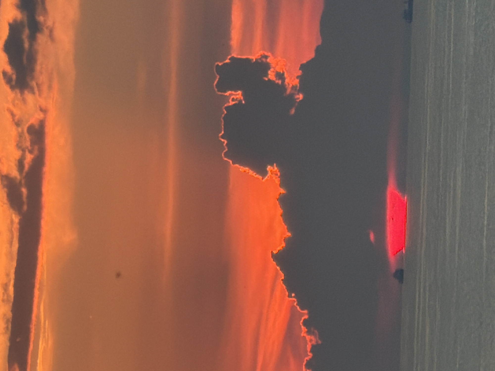 This is a vertical, eye-level shot of a vibrant sunset over the sea, taken from Sochi, Russia. The sky is a dramatic gradient of deep orange and red, with scattered dark clouds. One large cloud formation in the center is backlit by the setting sun, creating a bright, glowing outline. A portion of the sun, a bright red horizontal bar, is visible just above the water, partially obscured by a cloud. In the distance, on the horizon, two cargo ships are visible, one to the left and one to the right of the frame. The sea is calm with gentle waves reflecting the colors of the sky.