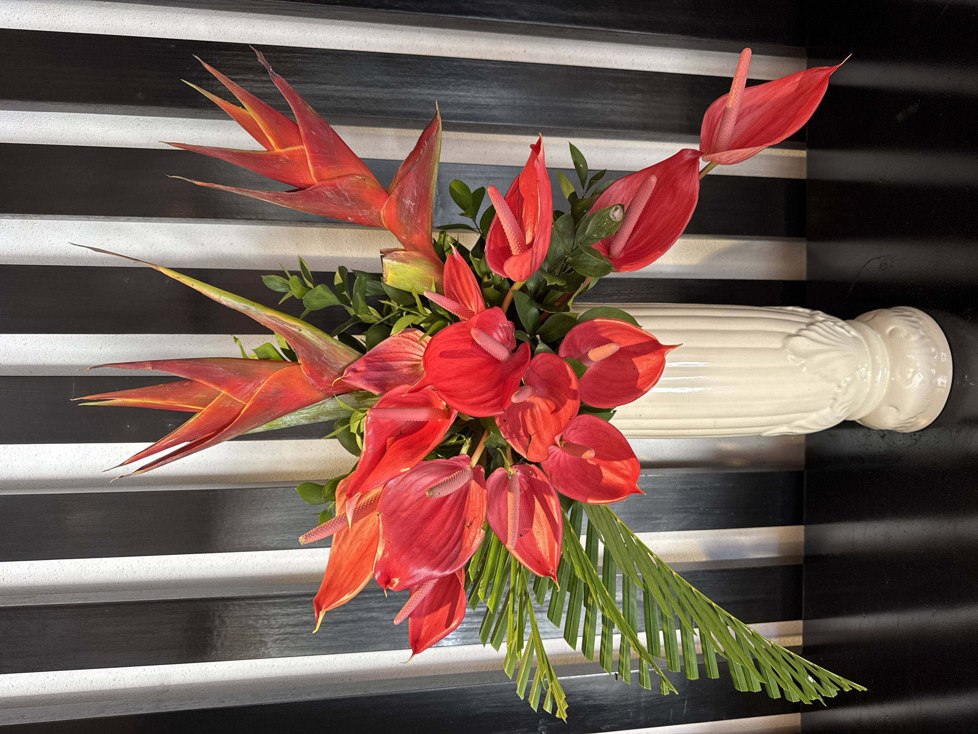 A top-down shot shows a vase of red and orange flowers placed on a surface composed of alternating black and white stripes. The flowers include red anthuriums, orange bromeliads, and green foliage. A white vase is partially visible on the right side. The image was taken in Kuta Selatan, Indonesia.