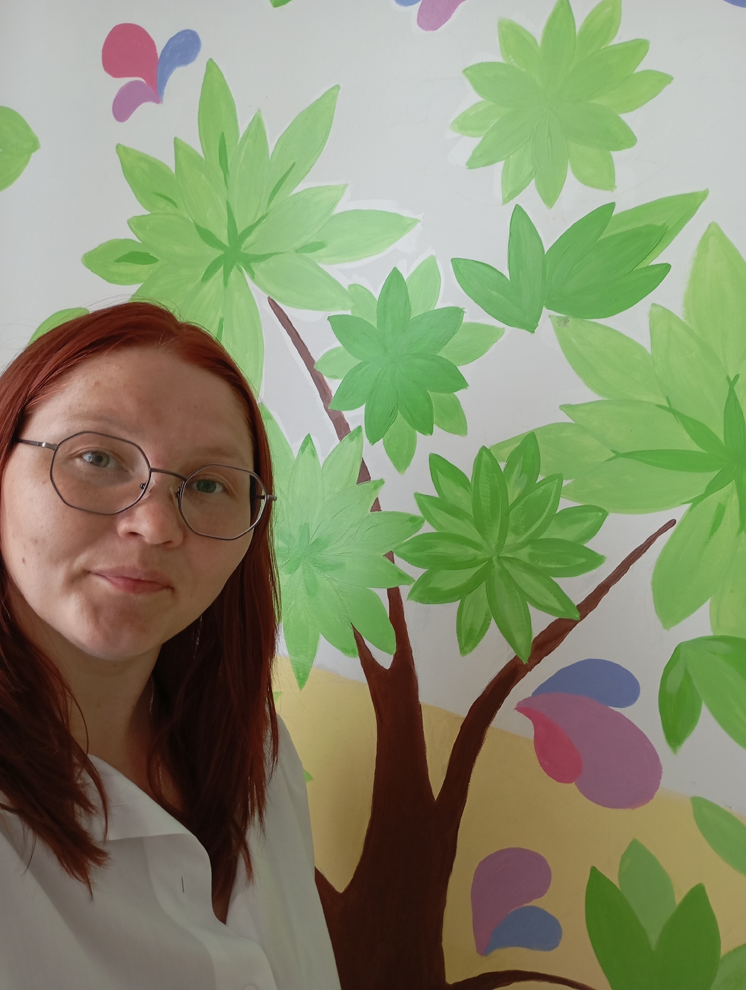 A person with red hair and glasses stands in front of a wall painted with a tree motif. The tree has a brown trunk and green leaves, with some pink and purple butterfly-like shapes painted around it. The person is wearing a white shirt. The location appears to be indoors, in a city and region identified as "Unknown, Unknown."