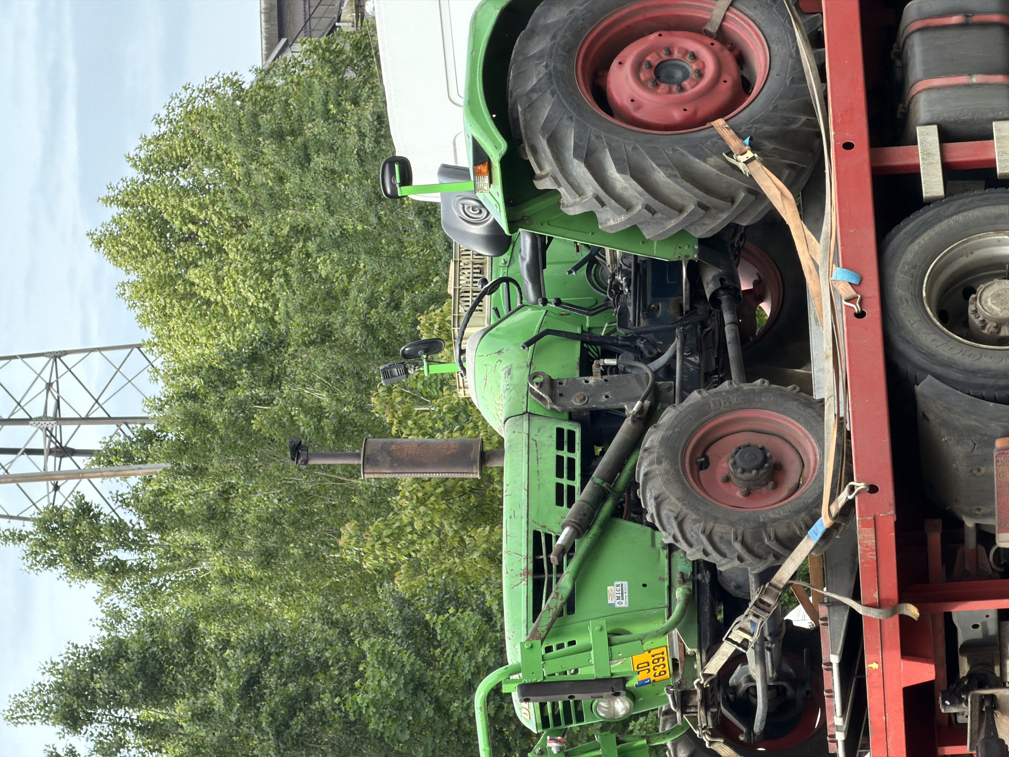 A green Deutz-Fahr tractor, license plate JD 6391, is secured on a red trailer. The location appears to be outdoors in Esch-sur-Alzette, Luxembourg, based on the license plate. The tractor has red rims and black tires, and it is secured with tan straps. The background includes green trees, a power line tower, a building, and a white truck. The tractor is the primary focus of the image.