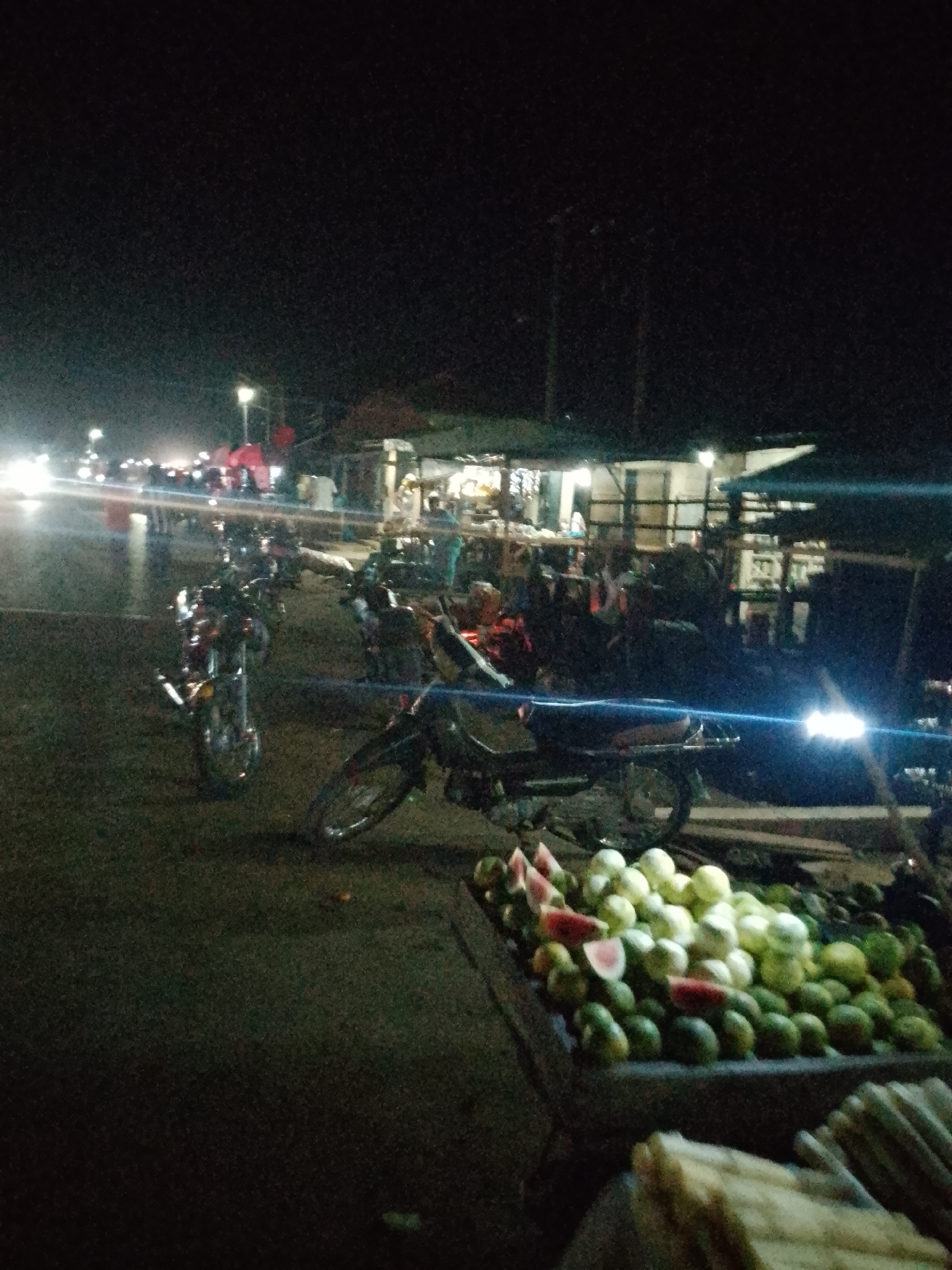 The image depicts a nighttime scene at a market stall in Serti, Nigeria. A variety of fruits, including watermelons and guavas, are displayed in a cart in the foreground. Several motorcycles are parked in the middle ground. In the background, a bustling market with stalls, lights, and moving vehicles suggests activity. The overall atmosphere is one of a lively nighttime marketplace.
