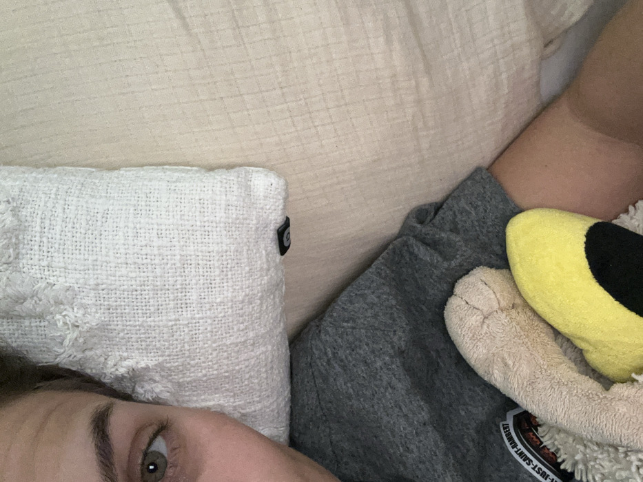 A person is partially visible, with one eye in focus. A white, textured pillow rests beside the person. A beige headboard or pillow is visible behind the white pillow. The person is wearing a gray garment, and a plush toy with yellow and beige fur is also present. The toy appears to have a sticker of some kind on it. The scene suggests a comfortable or relaxing environment, likely in a bed or resting space.