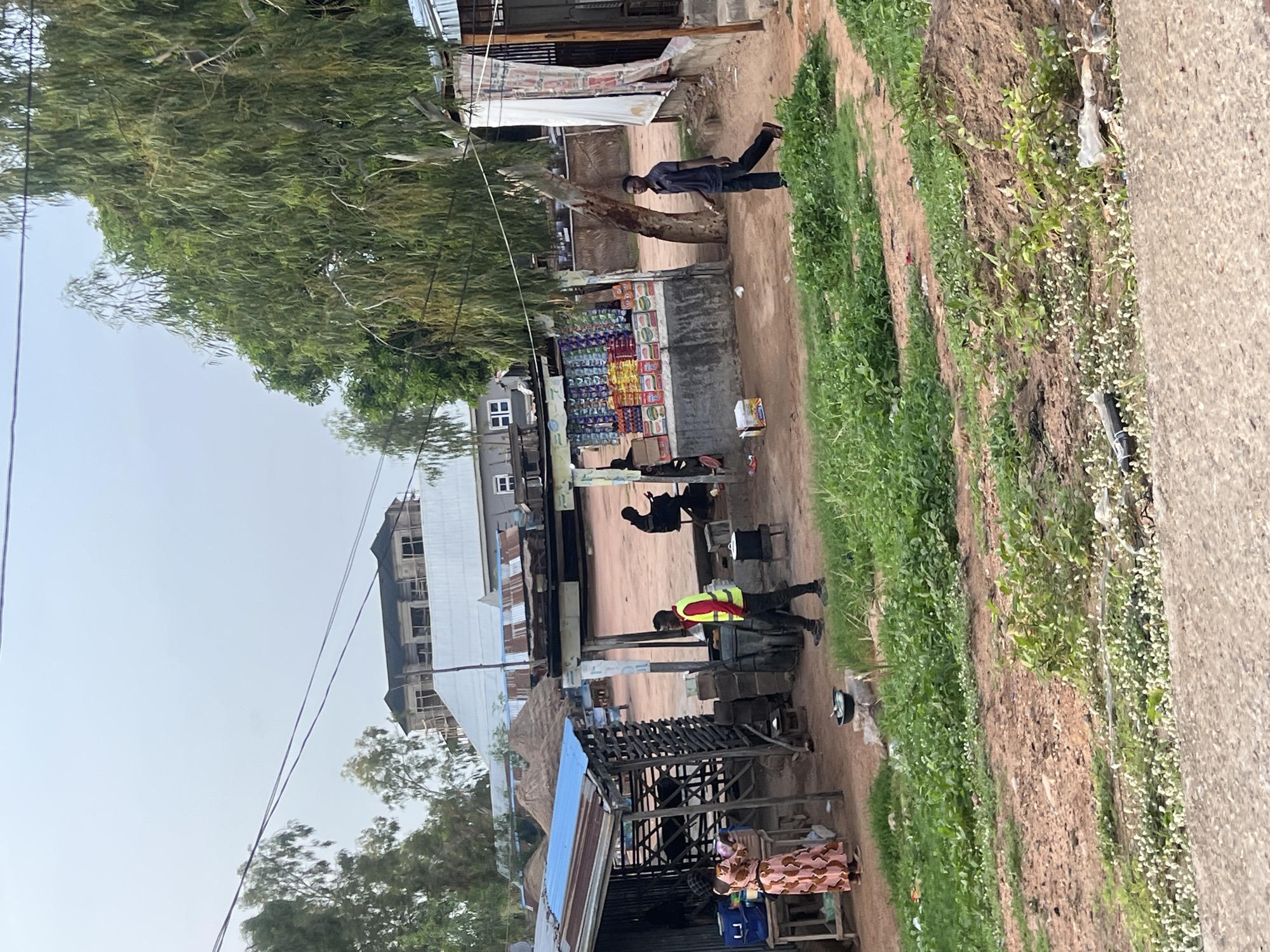 The image depicts a street scene in Kporva, Nigeria. The street is unpaved and bordered by grassy areas. Several individuals are present. One man stands in front of a small shop stocked with goods. Another man wearing a reflective vest appears to be working near a small structure. Further down the street, a person sits at what appears to be a workstation. Buildings of varying sizes and construction are visible in the background. Overhead power lines extend across the sky.