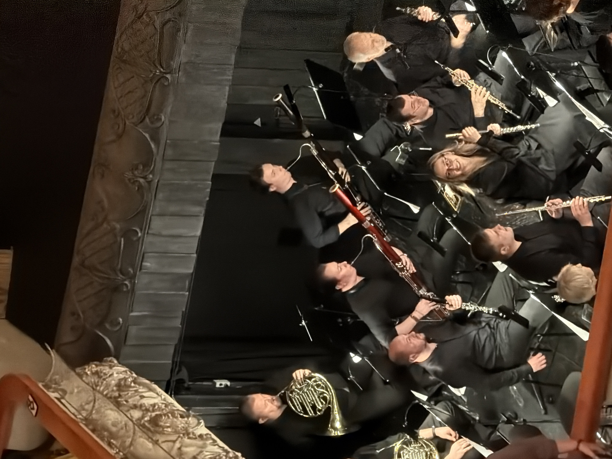 This image depicts an orchestra performing on a stage, likely in Lednevo, Russia. The orchestra members are dressed in black. In the foreground, on the left, a musician plays a French horn. To the right of the French horn player, a musician is playing a bassoon. Further to the right, another bassoon player is visible. In the middle ground, a man plays a clarinet, and to his right, a woman is playing a flute. Behind them, several other musicians are seated, holding various instruments, though their specific instruments are not clearly identifiable. The stage appears to be set against a dark curtain backdrop, with ornate detailing visible on the ceiling or proscenium arch above the stage, suggesting a formal performance venue.