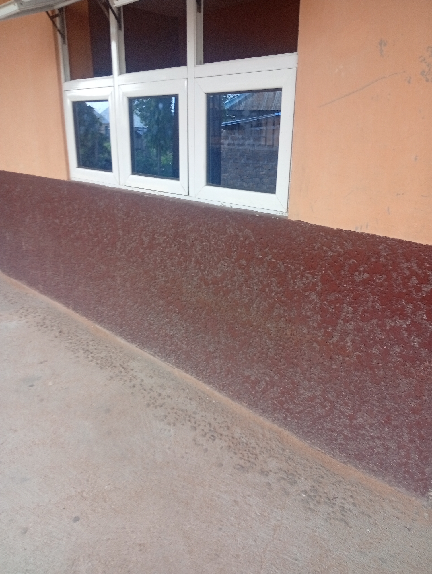 The image shows the exterior of a building in Jalingo, Nigeria. The building has a light orange-colored wall with a horizontal brown band running across the lower portion of the wall. The brown band appears to be textured, possibly stucco or a similar material. Above the brown band, there are white-framed windows with dark blue or tinted glass. The windows are divided into multiple panes, and some of them are open at an angle, allowing a view of trees and what appears to be other buildings or structures outside. The ground in front of the building is a light gray concrete surface, also textured, and it slopes downwards from right to left. The scene appears to be taken outdoors during the daytime, with natural lighting. No people or specific activities are visible in the image.