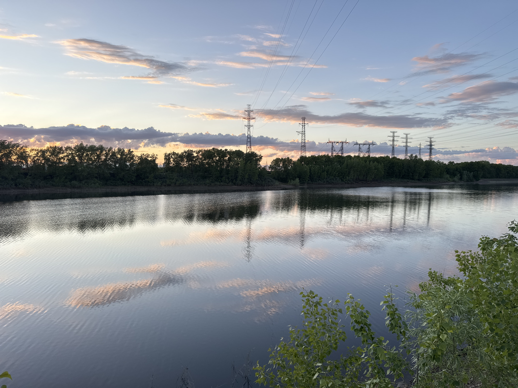 This media file depicts a tranquil waterscape at dusk in Chaikovsky, Russia. The scene features a wide body of water, likely a river, reflecting the colors of the twilight sky and the silhouettes of industrial structures.
In the background, a line of dense, dark green trees stretches across both banks of the water. Beyond the trees, numerous tall electrical transmission towers are visible, carrying a complex network of power lines that span the sky from left to right. The towers vary in design, with some having intricate lattice structures and others appearing more robust.
The sky is a prominent feature, displaying soft hues of light blue, lavender, and gentle orange-pink, particularly along the horizon, suggesting either a late evening sunset or an early morning sunrise. Scattered clouds, tinged with these warm colors, are spread across the sky.
The water surface is calm but shows ripples, particularly where the light from the sky and the reflections of the power lines and clouds create subtle patterns. The reflections on the water are clear, mirroring the sky's colors, the forms of the clouds, and the dark, angular shapes of the electrical towers and lines. Green foliage is visible in the immediate foreground on the right, partially obscuring the water's edge.
There is no visible human activity or specific event taking place, and no discernible text in the image. The overall atmosphere is one of serene natural beauty interspersed with industrial infrastructure during the quiet hours of twilight.