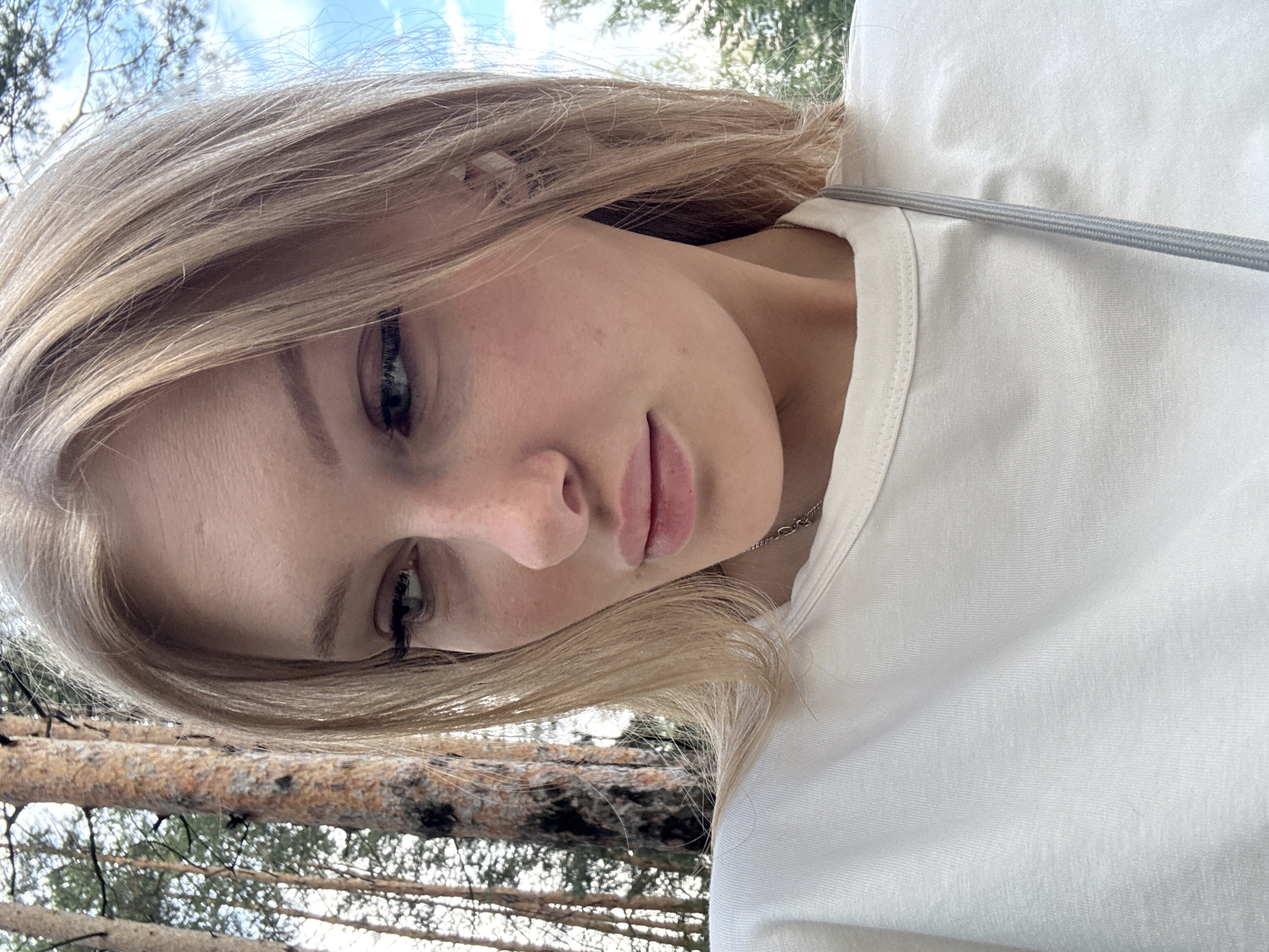 The image shows a young woman with blonde hair and fair skin, looking down and to her left. She is wearing a white t-shirt and a delicate silver necklace. She has noticeable freckles on her cheeks and a single earring in her visible ear. She is outdoors, with a blurred background of tall trees and a bright blue sky with some clouds. The trees suggest a forest or park setting. The lighting indicates it is daytime. There are no specific landmarks or elements visible that would definitively place this scene within the city of Perm, Russia. The image is a close-up, focusing on the woman's face and upper body.
