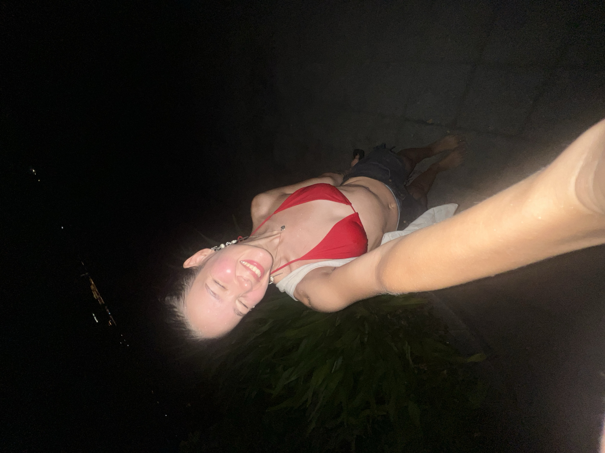 A woman is lying on her back wearing a red bikini top and dark shorts. She has a white cloth beneath her torso. The environment is dark, with foliage visible near her head and feet. A person's arm is visible, seemingly holding the camera to take a selfie. The location is potentially Nha Trang Ward, Vietnam.