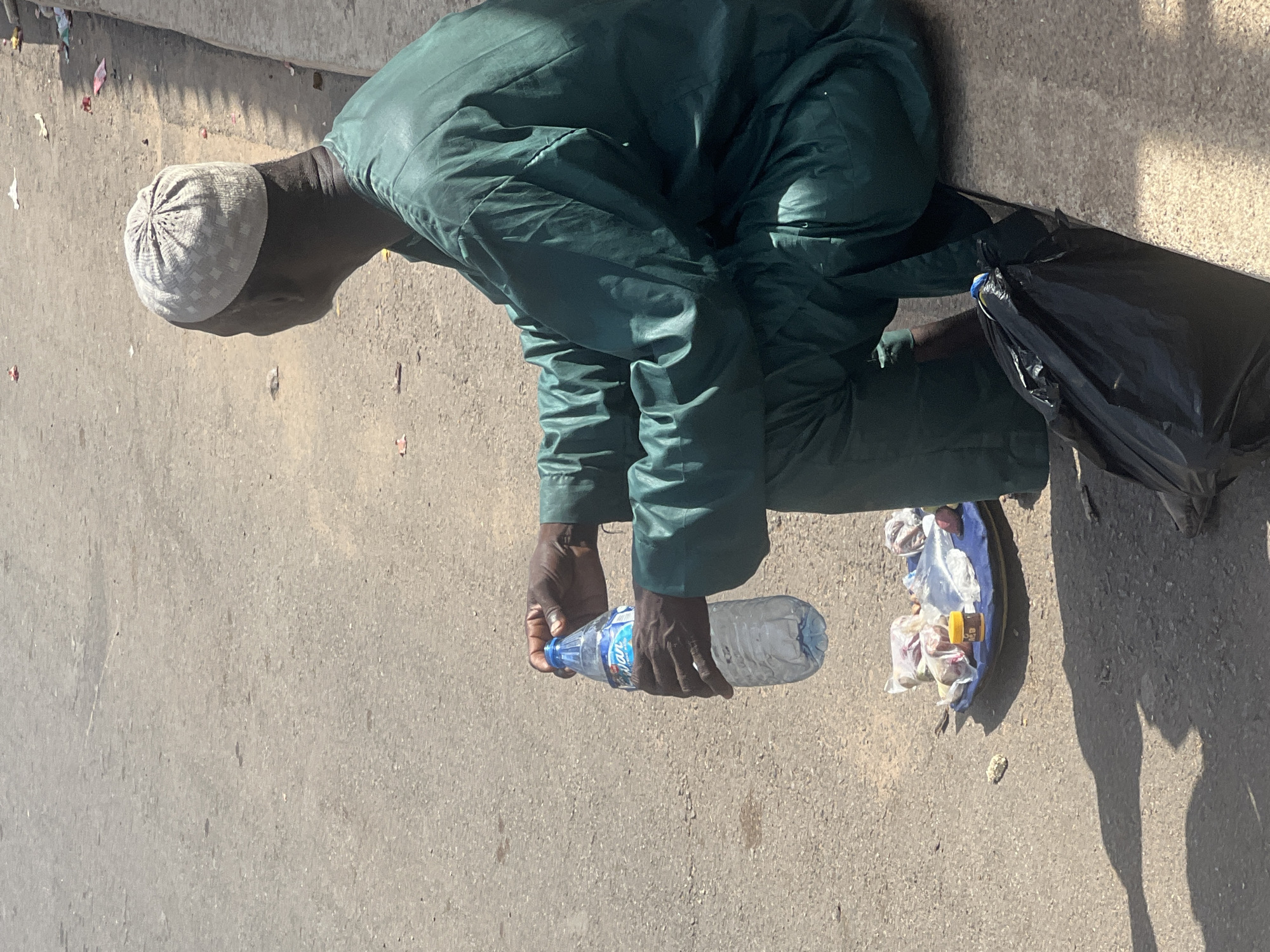 A man wearing a green outfit and a white kufi is kneeling on a concrete surface, holding a plastic water bottle. Next to him is a black plastic bag and a blue tray containing packaged items. The scene appears to be outdoors during the daytime, likely in a public space within the city of Jos, Nigeria. The setting includes scattered debris on the ground.
