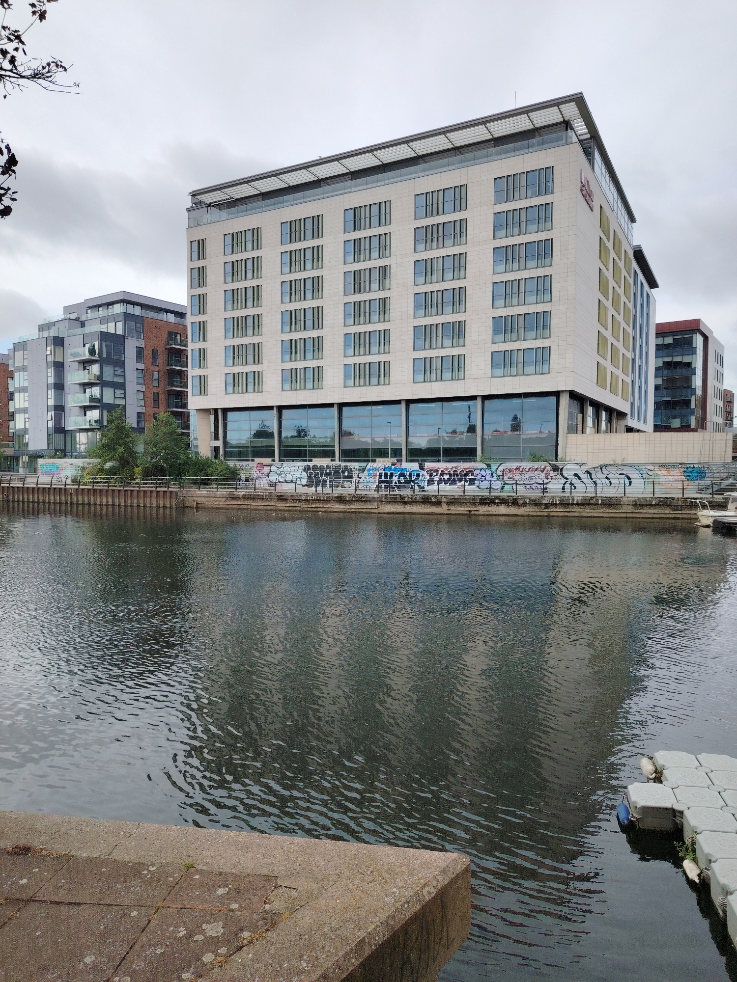 This image depicts an urban waterside scene in Peterborough, United Kingdom, specifically along the River Nene in the Fletton Quays area.
In the center of the frame, a multi-story building with a light beige facade dominates the view. This building, identified by a visible "Hilton Garden Inn" logo on its upper right side, features numerous regularly spaced rectangular windows across its front and side, some with vertical louvres or sunshades. The building has a flat roof with a visible rooftop structure and glass railings.
Below the main building, a section of the wall bordering the water is covered in multi-colored graffiti tags and lettering. A metal fence runs along the top of this wall. Behind the wall, a small cluster of green trees and shrubbery is visible.
To the left and behind the Hilton Garden Inn, several other modern buildings are present. These structures vary in height and feature combinations of dark grey and brick facades with numerous windows and balconies. To the right of the Hilton Garden Inn, another multi-story building with a reddish-brown upper section and dark glass is partially visible.
The foreground is dominated by a body of dark, slightly rippled water, identified as the River Nene. Reflections of the buildings, particularly the Hilton Garden Inn, and the overcast sky are discernible on the water's surface. In the bottom left, a concrete quay or pathway forms a dark grey and beige angular edge. In the bottom right, a modular floating platform composed of light grey square pontoons extends into the water, with a small white boat docked nearby.
The sky is overcast with a uniform light grey cloud cover. In the upper left corner, dark, bare tree branches extend into the frame. No individuals are visible in the scene.