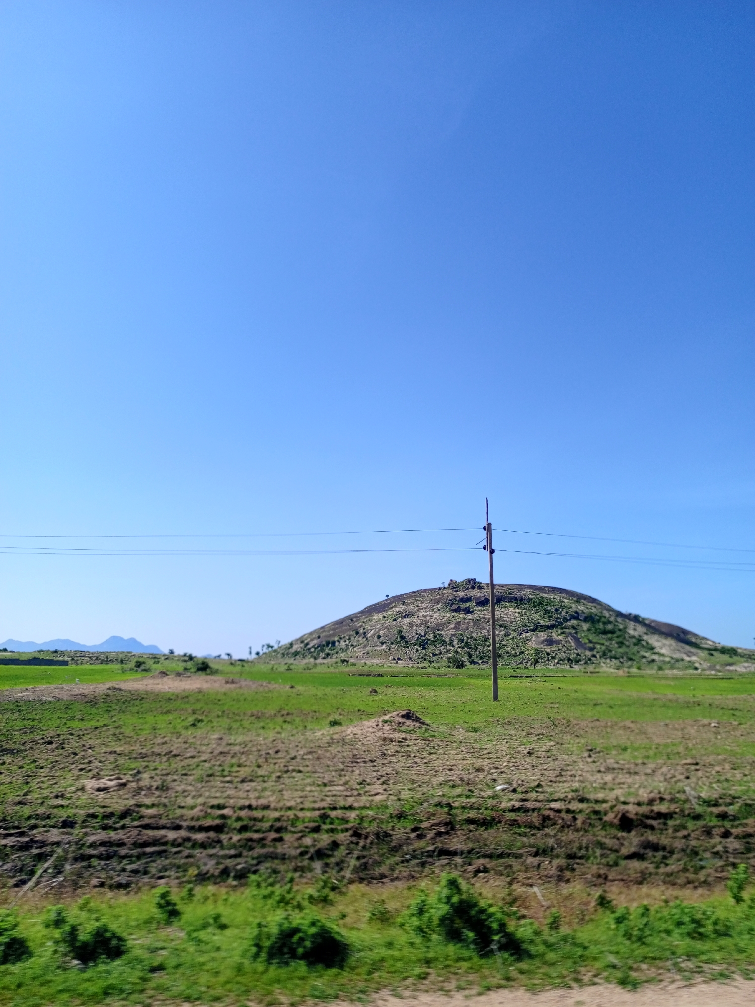 This is an outdoor, daylight, eye-level shot of a vast, grassy plain under a clear blue sky. In the mid-ground, a rounded, rocky hill dominates the landscape, its slopes covered in sparse green vegetation. A single wooden utility pole stands in front of the hill, supporting two thin wires that stretch horizontally across the frame. The foreground consists of a field of dry, brown earth with furrows, bordered by lush green grass in the very front. In the far distance, faint outlines of mountains can be seen on the horizon. There are no people or animals visible in the image. The scene appears to be a rural area, possibly on the outskirts of Bauchi, Nigeria.