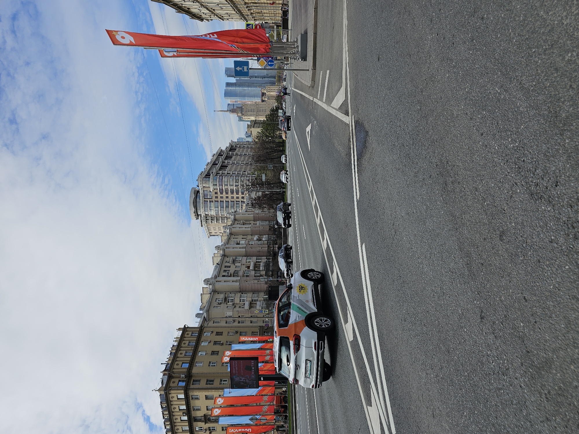 The image depicts a street in Moscow, Russia. The view is from a slightly elevated position, looking down the street towards a cluster of modern high-rise buildings in the distance. The sky is blue with scattered clouds. Along the left side of the street, there are older, Soviet-era buildings and a series of red banners or flags attached to the buildings. On the right side of the road, there is a sidewalk, and then the multilane road. Several cars are parked along the road, including one white car with orange and green branding. The Moscow City skyscrapers are visible in the distance, suggesting the street is located in the proximity of the Moscow International Business Center (Moscow City).
