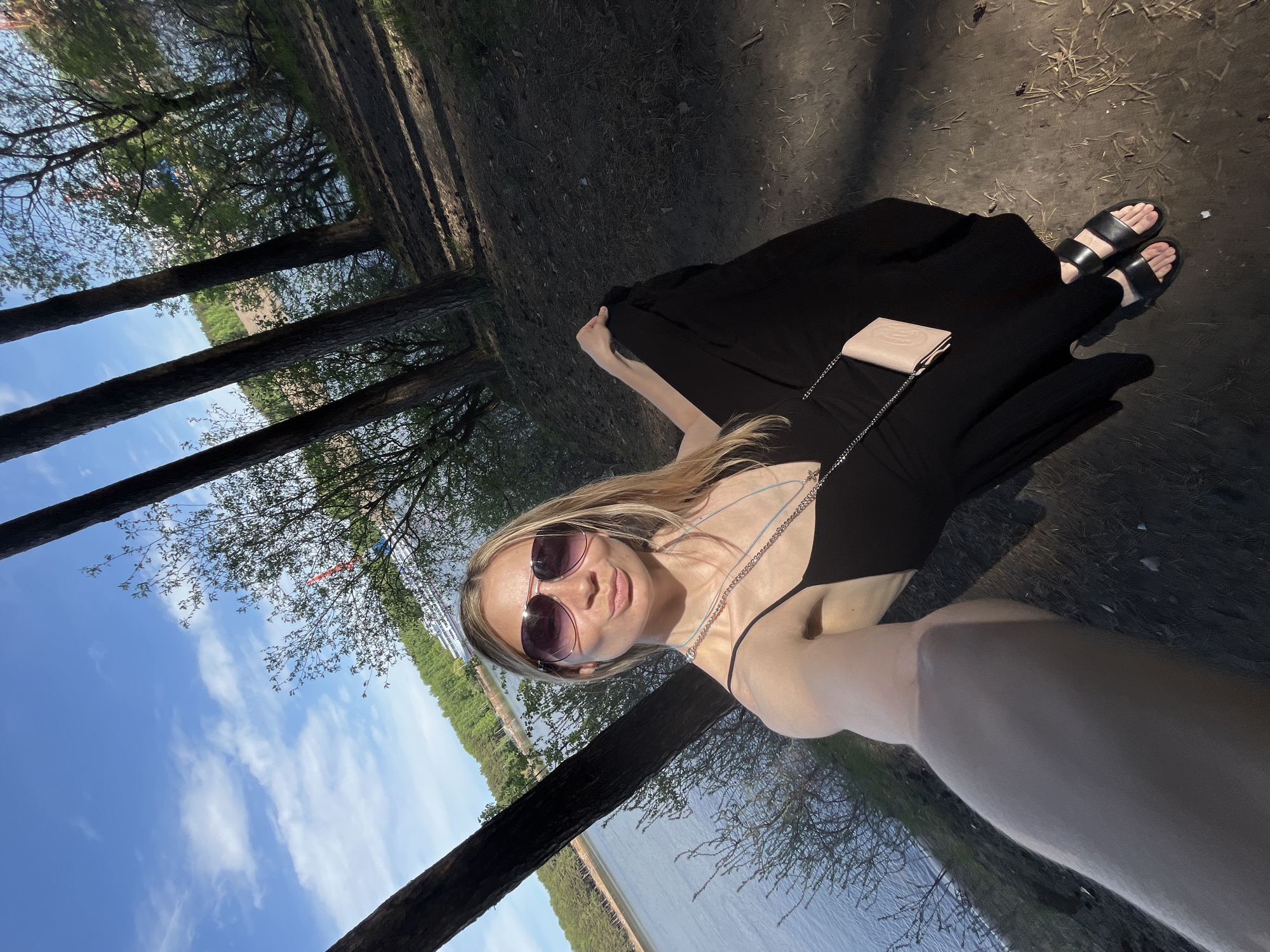 A young, fair-skinned woman with blonde hair and sunglasses poses for a selfie outdoors. She is wearing a black dress and sandals, with a small tan purse at her side. The photo appears to be taken in a wooded area next to a body of water. Trees frame the sky and water in the background. Based on the surroundings (a lake, the buildings visible through the trees, tree species) the photo may have been taken in the city of Chaikovsky, Russia. The ground is covered with dirt and fallen leaves.