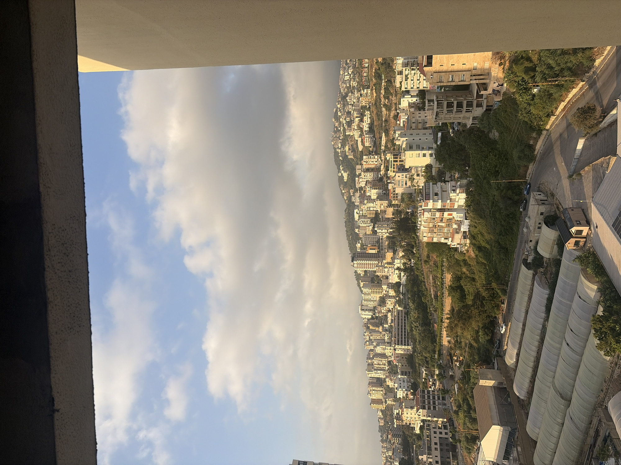 The image presents a panoramic view of a hillside city with a dense collection of buildings, suggestive of Wata Sillam, Lebanon. The city extends up a gentle slope, dotted with numerous residential and commercial structures. Many buildings are multi-story, with a variety of architectural styles, some appearing modern and others older. Lush green vegetation is interspersed throughout the urban landscape, particularly on the hillsides, indicating a natural environment integrated with the city. In the foreground, several long, tunnel-like structures, likely greenhouses or agricultural coverings, are visible. Roads wind through the scene, with a few cars present, suggesting active movement within the city. The sky is partly cloudy, with some large, diffused clouds obscuring parts of the sky and casting subtle shadows on the landscape, indicating it is likely late afternoon or early morning with the sun illuminating the buildings from the side. The overall impression is a bustling, lived-in urban environment situated within a verdant, hilly terrain.