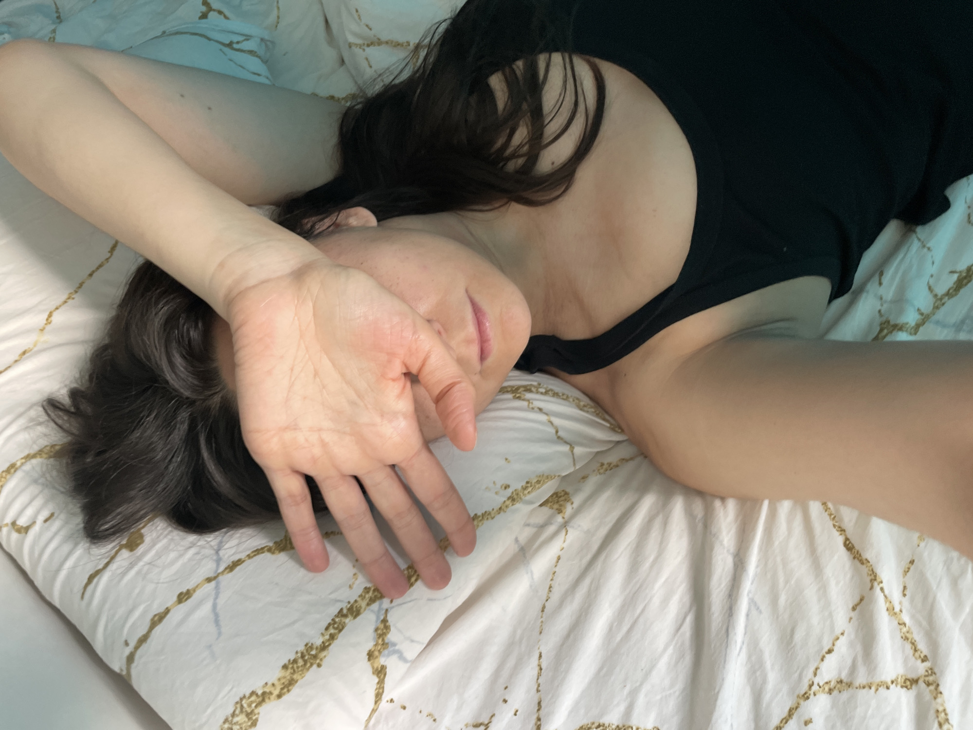A single individual, appearing to be an adult female with dark hair, is depicted lying on a bed in a relaxed posture. Her head is positioned on a white pillow with an irregular gold-and-blue abstract line pattern. She is wearing a black tank top. Her right hand is placed over her face, covering her eyes and upper nose, with the palm oriented towards the camera. Her lips are slightly parted, revealing a pink interior. Portions of her arms are visible, resting on the patterned bedding. The bed is covered with white sheets featuring the same gold and blue abstract line pattern. The environment is an indoor setting, likely a bedroom, with soft, diffused lighting. The specific location is the city of Unknown, Unknown.