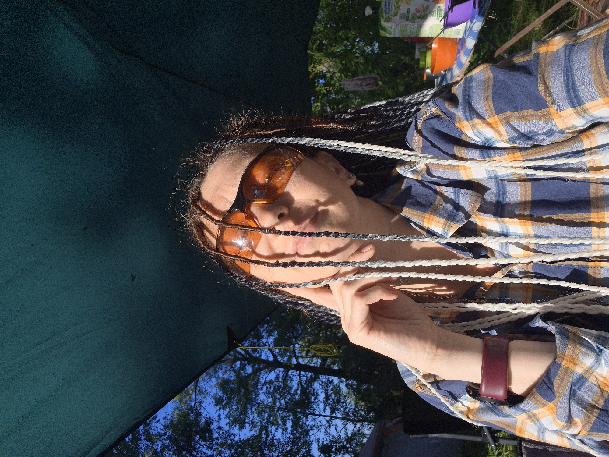 A medium shot captures a person with braided hair, wearing orange-tinted sunglasses and a plaid shirt, under a dark green canopy. The person's right hand is raised, with fingers touching their temple. The background shows trees and a blue sky. A table with various items, including a juice box and colorful containers, is visible to the right. The scene appears to be outdoors, possibly in a park or campsite.