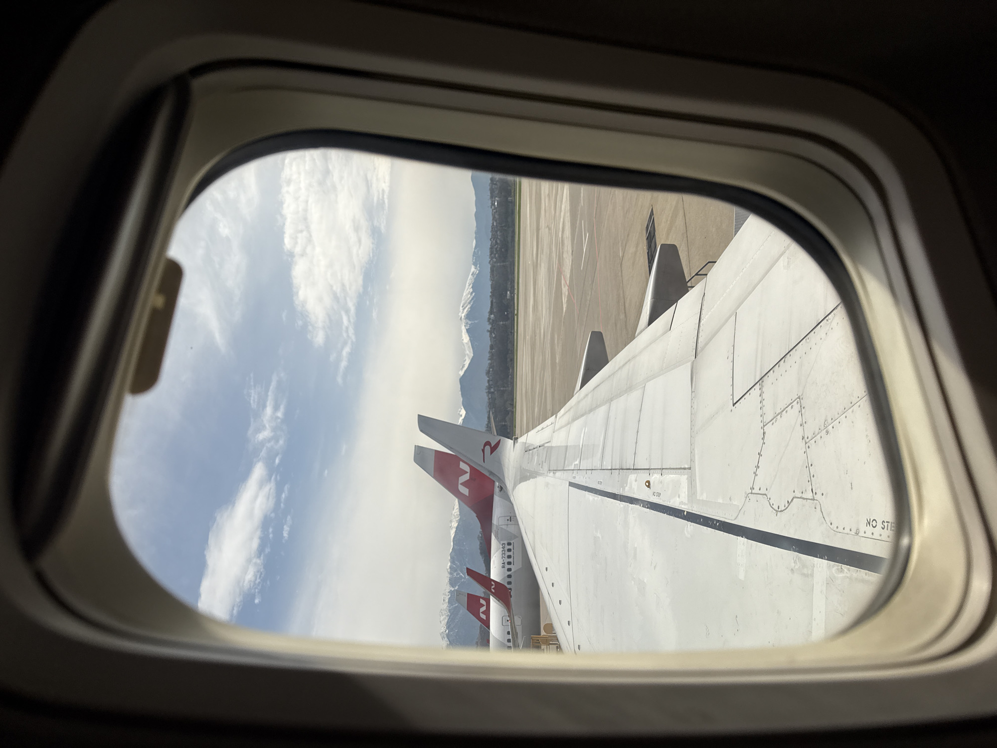The image shows a view from an airplane window. Outside the window, part of the wing of the airplane is visible, with other airplanes in the background, featuring tail fins with a stylized "2" in red. Beyond the airplanes is an airport tarmac and then a city skyline with mountains in the distance. Based on the aircraft tail number RA-73343 the image location can be identified as Sochi, Russia. The sky is partly cloudy.