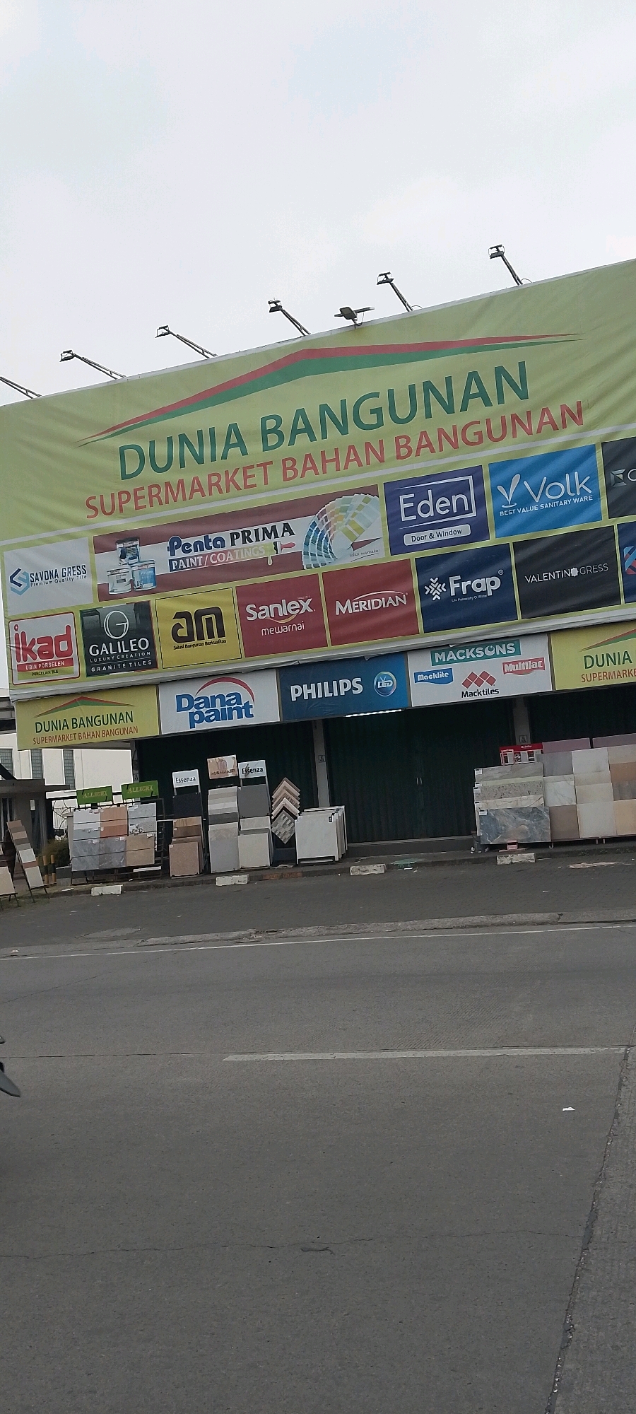 The image shows a street view in Cileungsi, Indonesia, featuring a prominent billboard for "Dunia Bangunan Supermarket Bahan Bangunan." The billboard, predominantly yellow and green, displays various brand logos associated with building materials, including "Eden," "Volk," "Penta PRIMA PAINT/COATINGS," "Sanlex," "MERIDIAN," "Frap," "VALENTINOGRESS," "ikad," "GALILEO GRANITE TILES," "an," "Macktiles," and "Multiplac." Below the billboard, a row of shops is visible, with displays of building materials like tiles and marble stacks placed outside. The street in the foreground is asphalt, with white lane markings and a sidewalk. The sky is overcast.