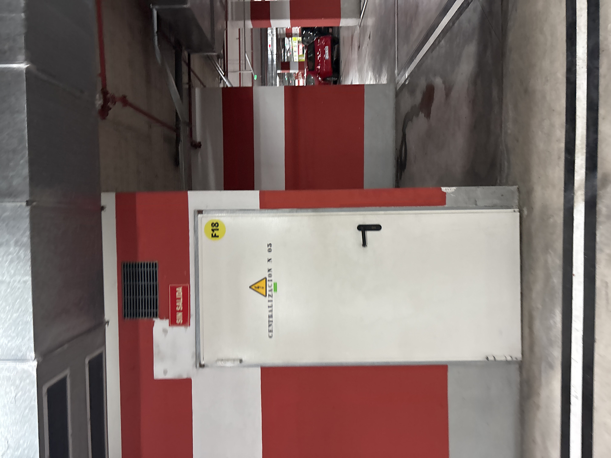 The image shows a door with the text "SIN SALIDA" which translates to "No Exit" on a red sign above it. There is also a yellow circle with "F18" on it. Below the yellow circle, the door has a symbol of a warning triangle with an exclamation mark inside, and text that reads "CENTRALIZACION N 05". The door has a black handle. The environment appears to be an indoor parking garage with red and white painted columns and a concrete floor. In the background, a red car is visible. The location is identified as Orihuela, Spain.