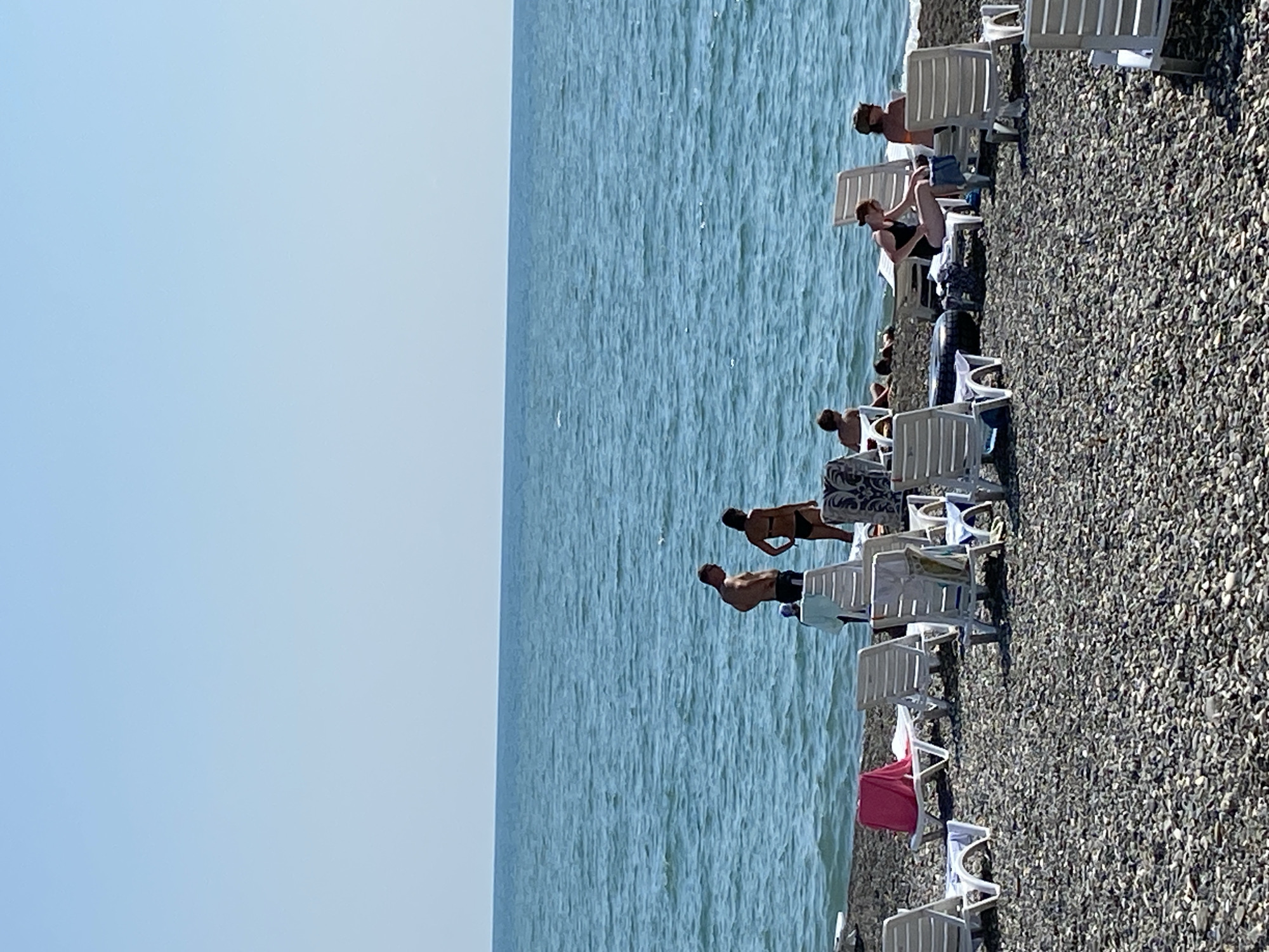 The image depicts a pebble beach in Chabkwara, Abkhazia, with the blue sea and clear sky in the background. Several white lounge chairs are scattered across the beach, some occupied by people. In the middle ground, a couple stands near the water's edge, with a woman in a bikini and a man in swimwear. Further down the beach, other individuals are seen relaxing on lounge chairs or sitting on the pebbles. A pink towel is draped over one of the lounge chairs. The overall scene portrays a tranquil beach environment on a sunny day.