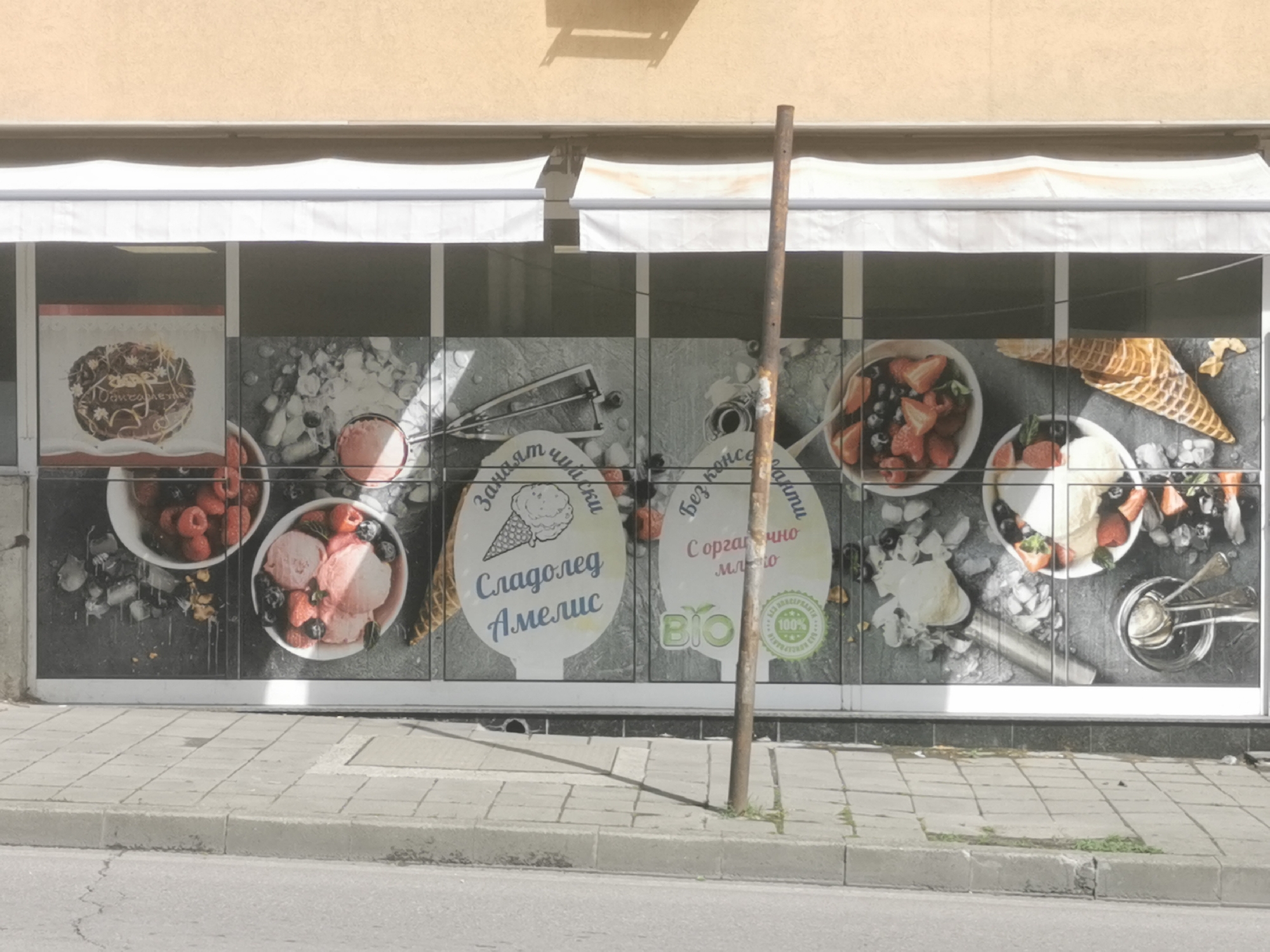 The image captures the storefront of a dessert or ice cream shop under bright daylight, likely in Blagoevgrad, Bulgaria. The upper part of the building shows a light yellow facade with two white retractable awnings, one of which has a faint orange stain. Below the awnings, the shop's large glass windows are covered with promotional graphics featuring various sweet treats.
The window display showcases appealing images of ice cream, fresh berries, and a dessert cake. On the left, a chocolate cake with frosting and a cherry is visible, along with bowls of berries (strawberries, raspberries, blueberries) and scoops of pink and white ice cream. Towards the right, more bowls of ice cream with berries and waffle cones are depicted, along with ice cream scoops and other dessert preparation tools. The graphics use a dark, textured background, making the colorful food items stand out.
Several text elements are incorporated into the window display, written in Bulgarian. A prominent label features an ice cream cone graphic and the text "Сладолед Амелис" (Sladoled Amelis), translating to "Amelis Ice Cream," preceded by "Запазена марка" (Zapasena marka), meaning "Registered trademark." Another text bubble states "без консерванти" (bez konservanti), which means "without preservatives." A third label reads "С органично мляко" (S organichno mlyako), indicating "With organic milk," and includes a "BIO" logo and a green circular emblem with "100%" and the word "Сертифицирано" (Sertifitsirano), suggesting it's certified organic.
In the foreground, a tall, rusty metal pole stands vertically, partially obscuring the view of the window graphics. The ground consists of a grey concrete sidewalk with a curb, adjacent to an asphalt road. A shadow of an unknown object falls onto the building facade above the awnings, and another shadow, likely from the metal pole, stretches across the sidewalk.