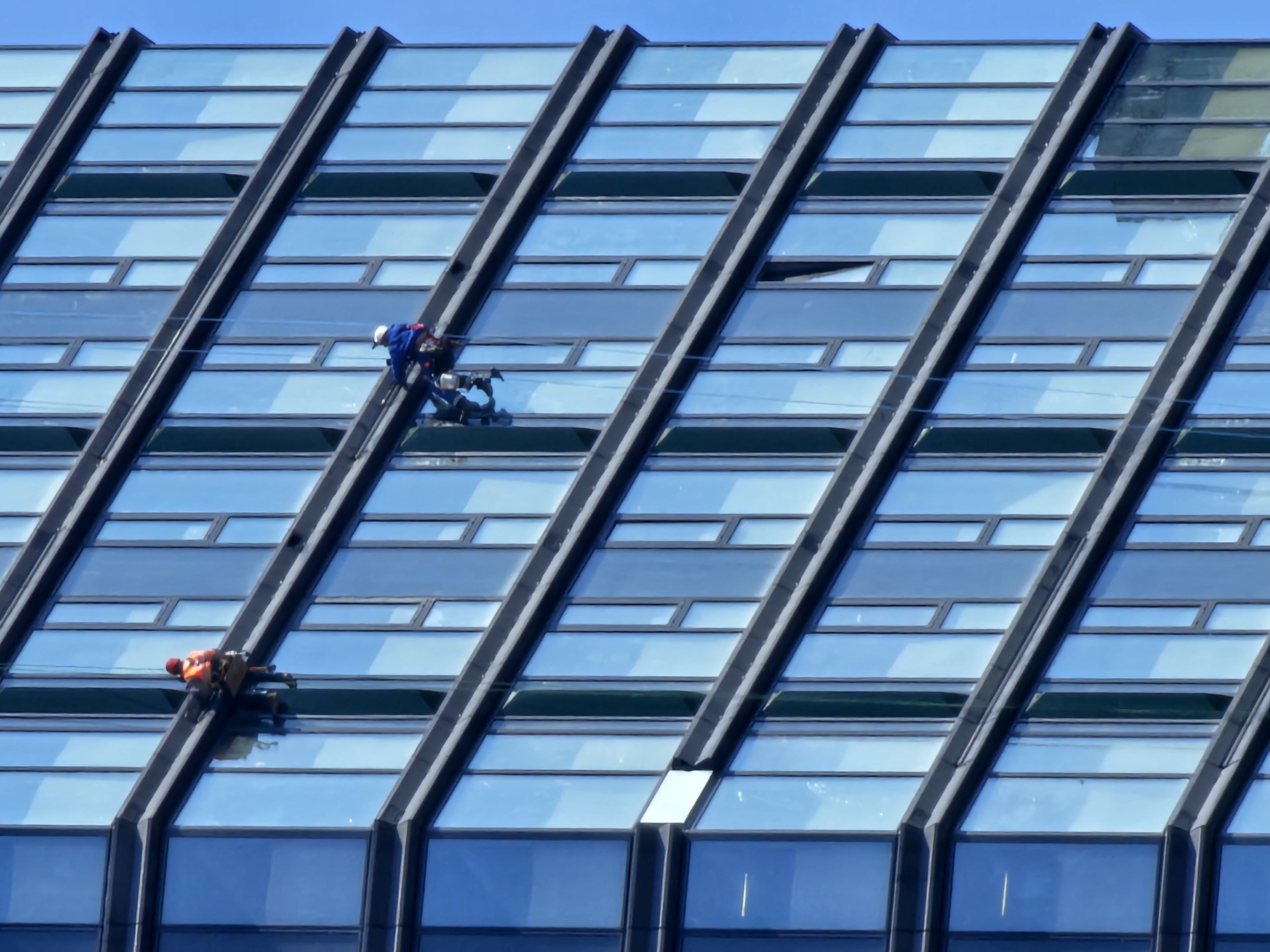 The image shows the exterior of a modern, high-rise building, likely an office or commercial structure, with a facade comprised of numerous blue-tinted glass panels arranged in a grid-like pattern. The architectural design includes prominent dark gray or black structural elements that create a diagonal linear pattern across the building's face.
Two window washers are visible, suspended by ropes and harnesses, performing maintenance on the building's glass exterior. One worker is wearing a blue jacket and a white hardhat, while the other is wearing an orange vest and a red hardhat.
The location in the city of Khimki, Russia, is only given and cannot be verified using visual data.