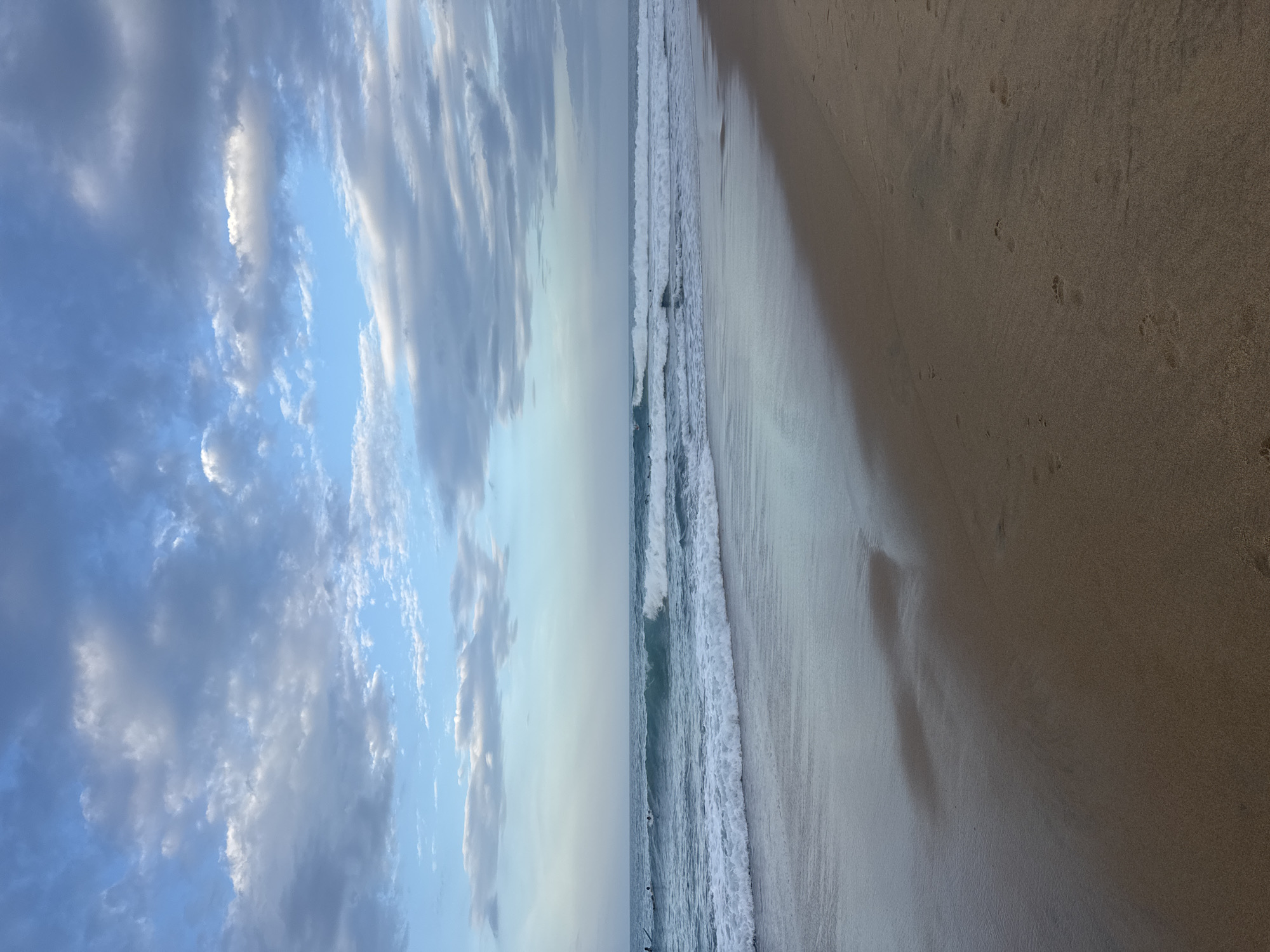 This media file captures a serene beach scene at what appears to be either sunrise or sunset, given the soft, diffused light and the color of the sky. The image is oriented vertically, presenting a panoramic view that divides the frame into distinct areas: the sky, the ocean, and the beach.
The left side of the image is dominated by a vast expanse of sky filled with dynamic clouds. These clouds are a mix of blues and whites, with some appearing wispy and others more solid, suggesting a dynamic atmosphere. The bright sunlight, though not directly visible, is filtering through these clouds, creating a luminous effect.
The middle section of the image depicts the ocean. Gentle waves are breaking along the shore, creating a foamy white line of surf. The water itself appears a tranquil blue-green. A few dark, indistinct shapes in the water suggest the presence of surfers or swimmers, engaged in activities like surfing or enjoying the water.
The right side of the image showcases a sandy beach. The sand is a light brown or beige color, with visible footprints, indicating recent human or animal activity. A shadow stretches across a portion of the sand, possibly cast by a person or an object not fully within the frame, adding depth to the scene.
The overall mood is peaceful and calming, evoking a sense of quietude and the natural beauty of a coastal environment. The location context, Arugam Bay, Sri Lanka, is known for its beautiful beaches and surfing spots, which aligns with the visual elements present in the image. There is no visible text in the media file.