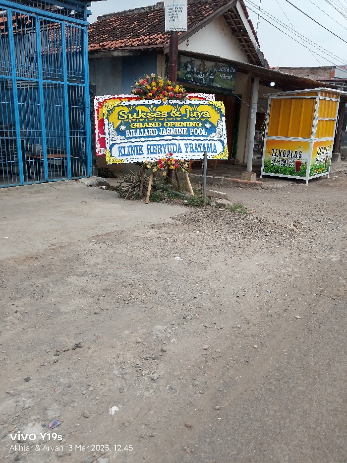 The image shows a street in Kecamatan Anjatan, Indonesia (latitude: -6.411, longitude: 107.961). There is a sign in front of a building that says "Sukses & Jaya Grand Opening Billiard Jasmine Pool Klinik Heryuda Pratama." The sign is decorated with flowers. There is a blue gate to the left of the sign and a yellow building with a "Lemarios" sign to the right of the sign. The ground is gravel. The image was taken with a mobile phone.