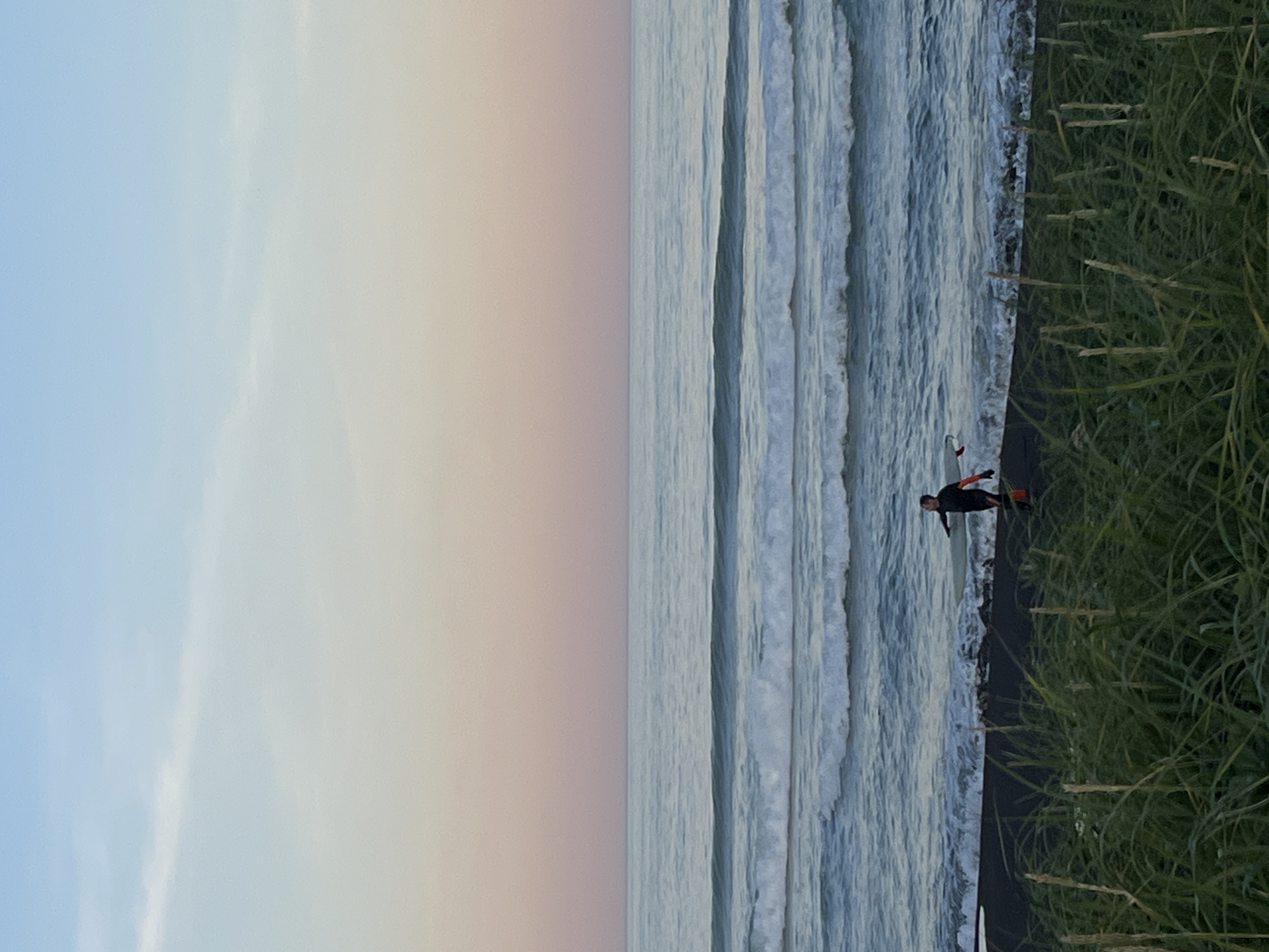In Petropavlovsk-Kamchatskiy, Russia, at dusk, a person wearing a black wetsuit with orange accents is standing on a black sand beach. The person is holding a red object, possibly a surfboard. Gentle waves are rolling in from the ocean, and there is a band of light green grass in the foreground. The sky above is a gradient of soft blue and pink.