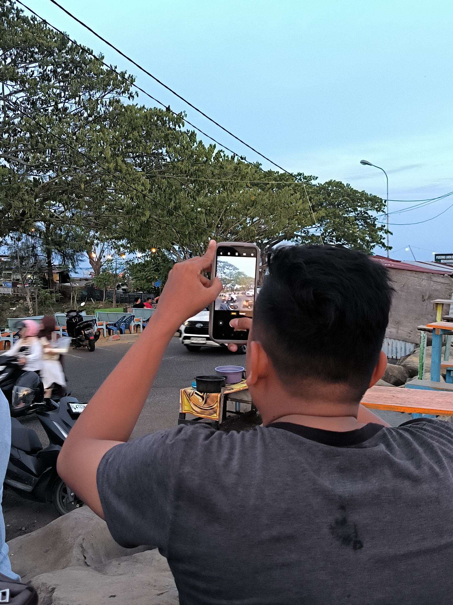 The image depicts a street scene in Lhokseumawe, Indonesia, likely during the evening. In the foreground, a person is holding up a smartphone, apparently taking a photograph of the street. The phone's screen displays a view of the road with traffic, including a white car and several motorcycles. The person's back and arm are visible, and they are wearing a dark grey t-shirt. Behind them, a busy street is visible with various vehicles and pedestrians. A large tree with dense foliage dominates the middle ground, casting shadows on the surroundings. To the right, there are some outdoor seating arrangements, possibly indicating a cafe or restaurant. In the background, buildings and utility poles are visible, and the sky is a pale blue, suggesting dusk. The overall atmosphere is that of a lively urban setting.