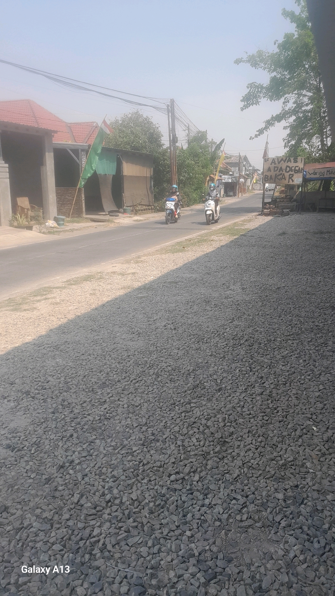 The image depicts a scene on a street in Kirig, Indonesia, likely during the daytime. The foreground is dominated by a vast expanse of grey gravel, casting a sharp shadow from the right side of the frame. A paved road runs horizontally across the mid-ground, with two individuals on motorcycles traveling away from the viewer. The rider on the left is on a white and blue motorcycle, while the rider on the right is on a white scooter. Both riders appear to be wearing helmets and casual clothing.
Along the side of the road, there are several structures. On the left, a building with a red-tiled roof and a grey facade is visible. Further down the road, a weathered brown structure with a corrugated metal roof is present, adorned with a green and white flag on a pole. To the right of the road, there's a sign with Indonesian text, "AWAS! ADA DEGAN BAKAR" (Caution! There is a burning coconut) and "Warung Makan" (Eating stall). More buildings and vegetation line the street in the distance. The sky is hazy and light blue, suggesting a warm or possibly polluted atmosphere. The overall impression is a functional, everyday street scene in a rural or semi-urban Indonesian setting.