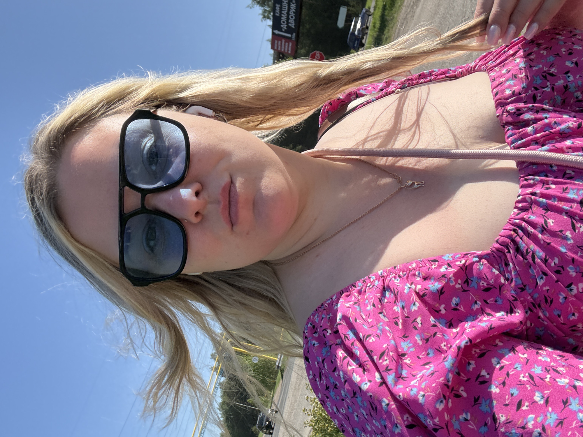 A fair-skinned woman with blonde hair is captured in a close-up, slightly angled portrait. She is wearing sunglasses with black frames, and an earbud is visible in her left ear. Her attire consists of a pink floral dress. She has a nose ring. A necklace with a bird pendant hangs around her neck.
The background features a street scene under a bright, sunny sky. A road, trees, and a sign with Cyrillic lettering are visible in the distance. The sign translates to "Cafe 'Homemade Yard'". A car is parked nearby. Based on the Cyrillic text on the sign, the location is identified as Noviy, Russia.