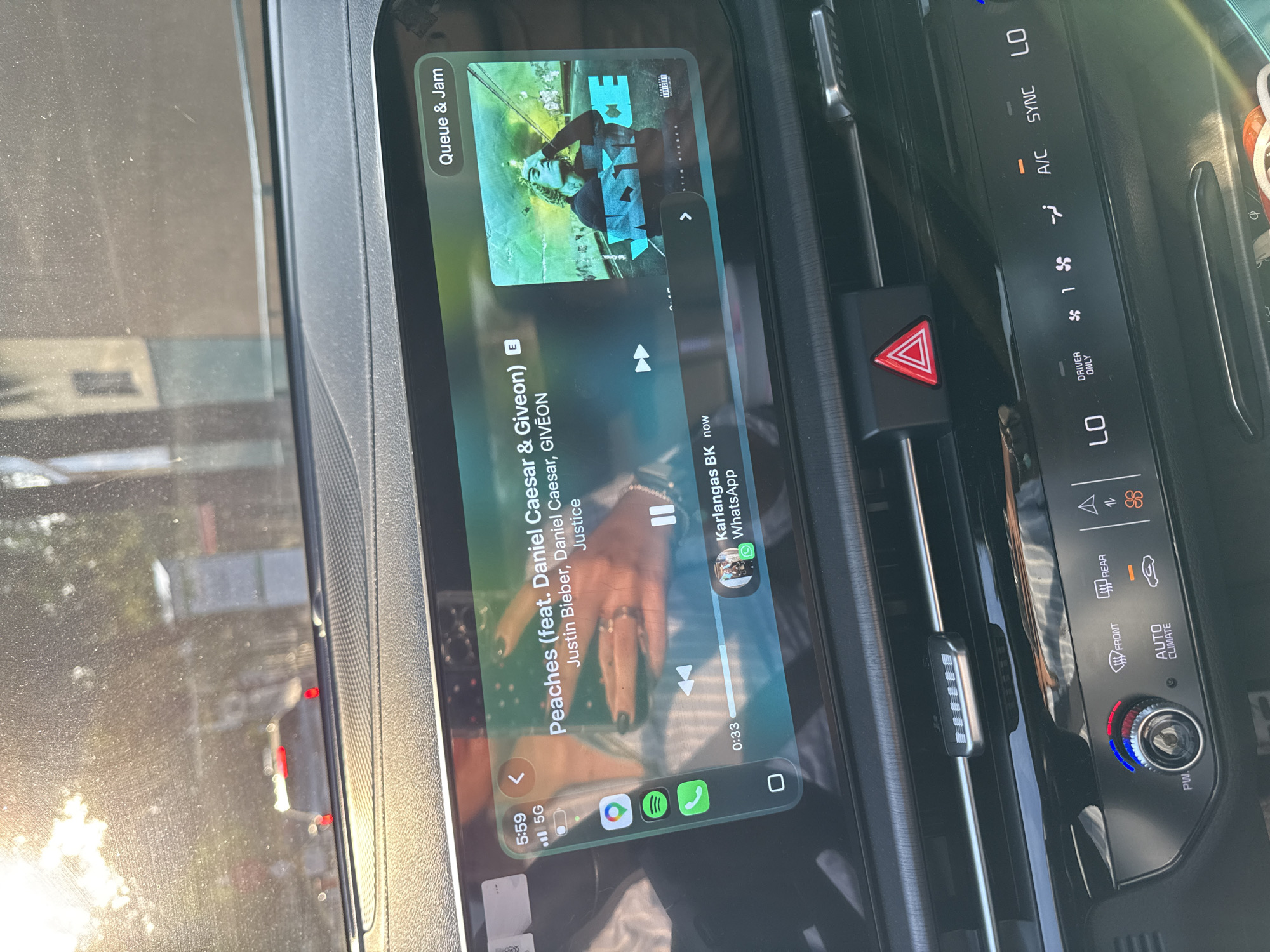 The image shows the interior of a car. The infotainment screen displays the song "Peaches" by Justin Bieber featuring Daniel Caesar & Giveon, playing on Spotify. There is a WhatsApp notification from Karlangas BK. The car's climate control panel is visible, with settings for A/C, front and rear defrosters, and AUTO CLIMATE, and LO indicators. The hazard light button is visible. The car appears to be in motion, with an out-of-focus view of the outside through the front window. The location in the city of San Bernardino Tlaxcalancingo, Mexico, is not discernible from the image.