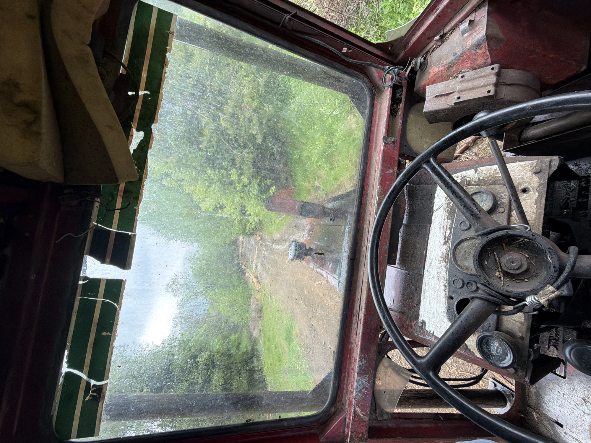 The image, taken from inside the rusted, worn cab of a vehicle in Рилски манастир, Bulgaria, features a view through the grimy windshield. The interior of the cab is decayed, with a black steering wheel dominating the right foreground and a damaged dashboard with gauges. Part of the interior is stained white. To the left of the windshield, remnants of a striped, possibly green and white, sun visor are visible. The view through the window reveals a dirt road leading into a lush green forest. There is a person standing some ways down the road, with another truck behind.