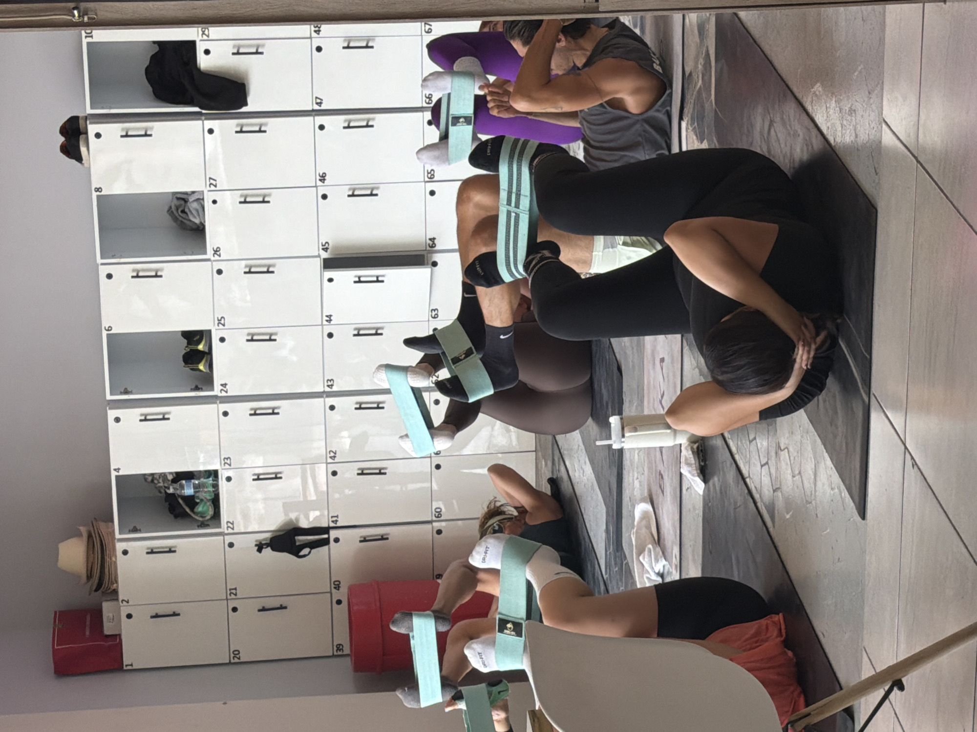 Here's a description of the image:
**Scene:** Interior view of a fitness studio or gym setting.
**People:** The image shows at least three individuals, likely in a fitness class. They are lying on exercise mats with their knees bent and feet on the floor. Each person has a resistance band looped around their thighs. One individual is in a traditional crunch position with their hands behind their head.
**Objects:** The primary objects present are the people performing the exercises, exercise mats, resistance bands, and a wall of lockers behind them. The lockers are white and numbered. Some lockers contain personal items.
**Actions:** The individuals are engaged in a core workout, focusing on abdominal exercises with the added resistance of the bands around their legs.
**Environment:** The studio has a tiled floor and good lighting. The lockers suggest this is a changing area connected to the workout space. The location in the city of Puebla, Mexico is not determinable from the image.