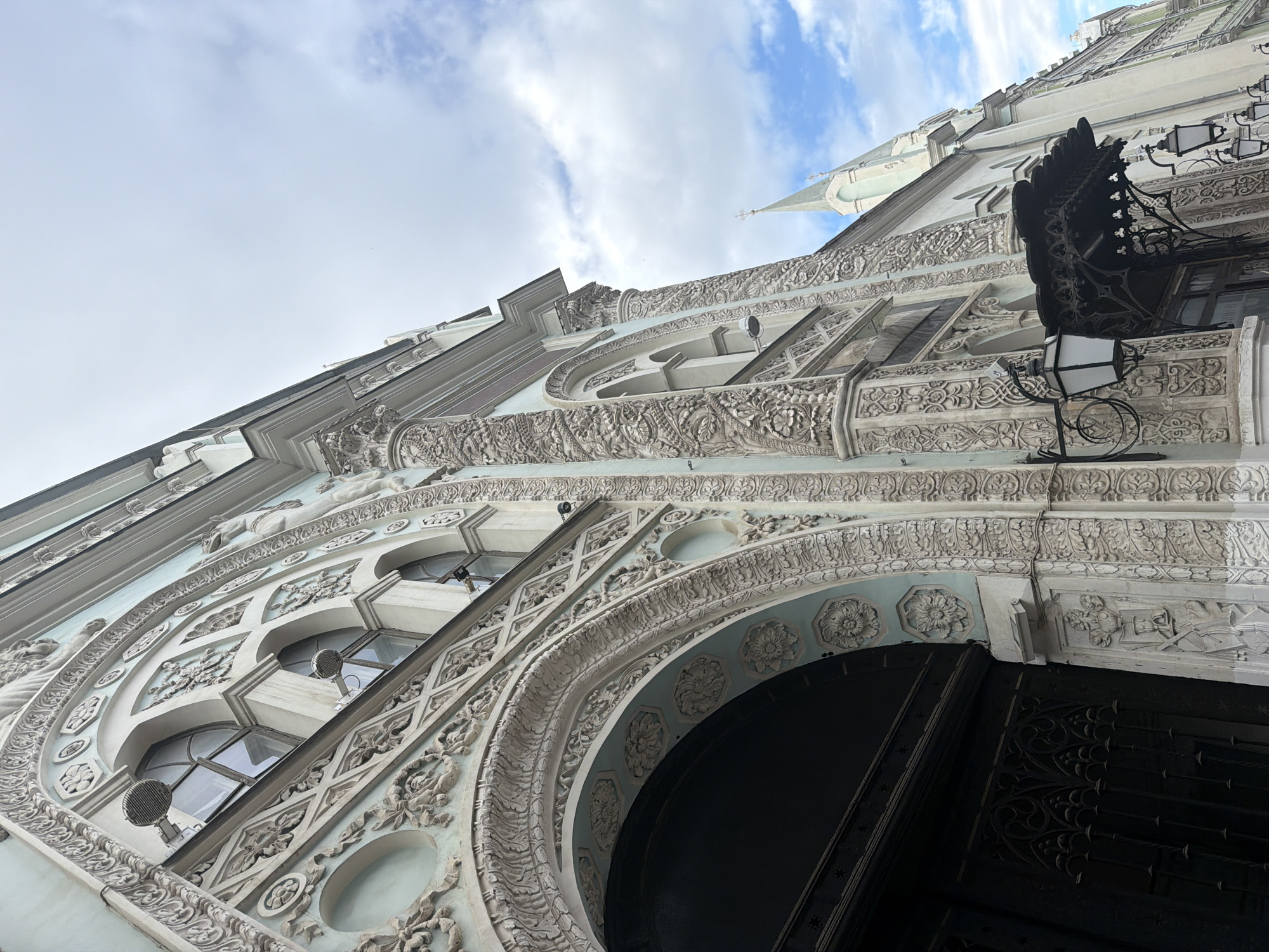 The image depicts a low-angle view of an ornate building facade in Moscow, Russia. The architecture is characterized by intricate relief carvings, arched windows, and decorative moldings. A tall, dark-colored arched doorway dominates the lower portion of the frame. Above, multiple stories feature rounded windows and detailed embellishments. A wrought iron lantern is affixed to the building's exterior on the right side. In the background, a spire is visible against a cloudy sky. The overall color palette consists of muted tones of white, gray, and blue.