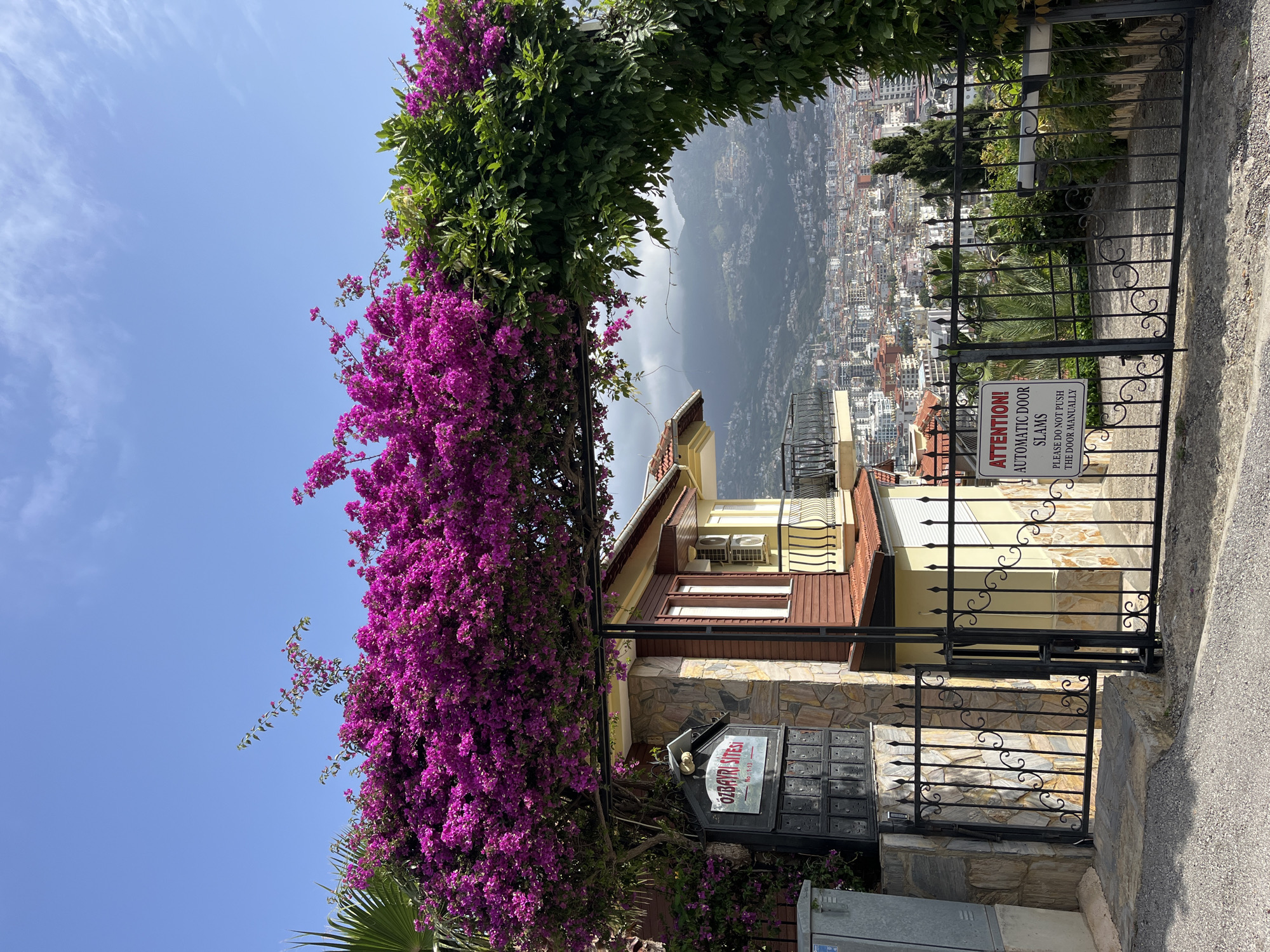 The image captures a residential scene in Alanya, Turkey. A wrought-iron gate stands in the foreground, bearing a sign that reads "ATTENTION! AUTOMATIC DOOR SLAMS. PLEASE DO NOT PUSH THE DOOR MANUALLY" and a mailbox indicating "ÖZBAYRI SITESI No: 1-13".
A building with a combination of stone and wood facade is visible behind the gate. Lush purple bougainvillea flowers drape over a structure, partially framing the scene. In the background, a cityscape of Alanya stretches out towards distant mountains, suggesting a hillside or elevated location. The sky is clear blue.
