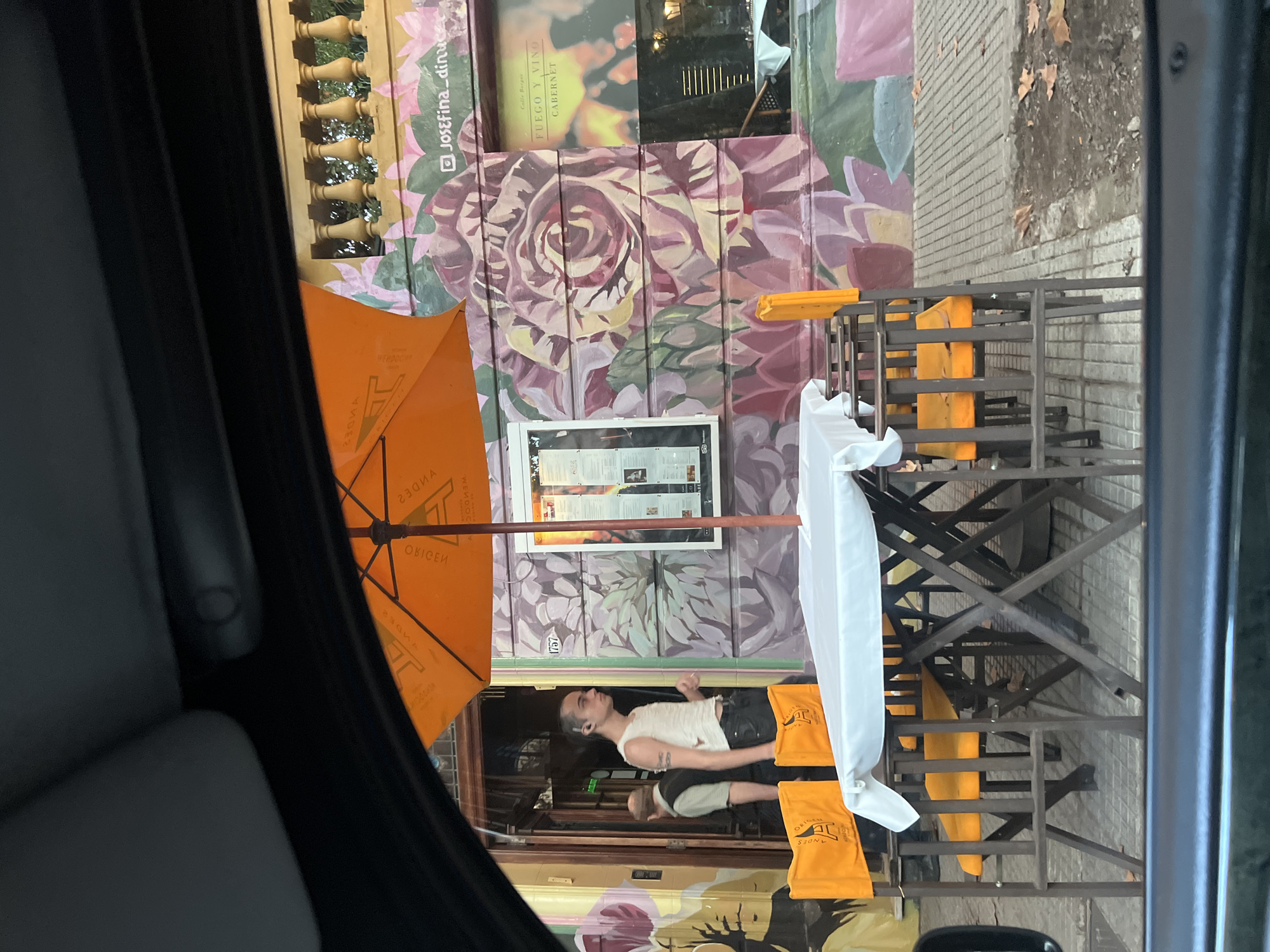 The image, taken from inside a vehicle, captures an outdoor seating area of a restaurant in Buenos Aires, Argentina. A man with a shaved head and tattoos on his arm lies on a restaurant chair. The restaurant façade features a mural with large floral designs in shades of purple, pink, and green, with a sign that reads "Josefina dinuc" and another sign mentioning "Calle Borges, FUEGO Y VINO CABERNET." The outdoor seating includes metal chairs with orange cushions and a large orange umbrella branded with "ANDES ORIGEN." The image shows a portion of a sidewalk and the outside wall of the restaurant. The camera is inside a vehicle, with part of the car's interior (seat, dashboard) visible.