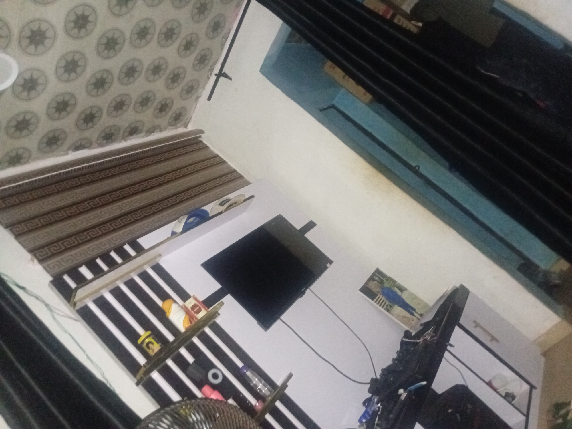 This image displays the interior of a domestic room in Ogbogbo, Nigeria, captured from an elevated and tilted perspective. The room features a white wall with black horizontal stripes, upon which two narrow wooden shelves are mounted. The top shelf contains a yellow container, an orange/yellow object, and a small package. The lower shelf holds a pink cup, a white ring-shaped object, and a clear plastic bottle. A black flat-screen television is mounted below the shelves on the striped wall, with visible cabling extending downwards. To the right, brown roller blinds adorned with a repeating geometric pattern are visible. The ceiling is covered with light-toned tiles, each featuring a central dark, star-like circular motif. In the lower right, a section of a dark blue wall or trim is present, alongside black vertical blinds. The lower left area includes a dark-topped, light-sided dresser or desk, cluttered with electronic components and wires. A framed picture, depicting a person in blue attire, leans against the wall on this surface. No individuals are present within the scene.