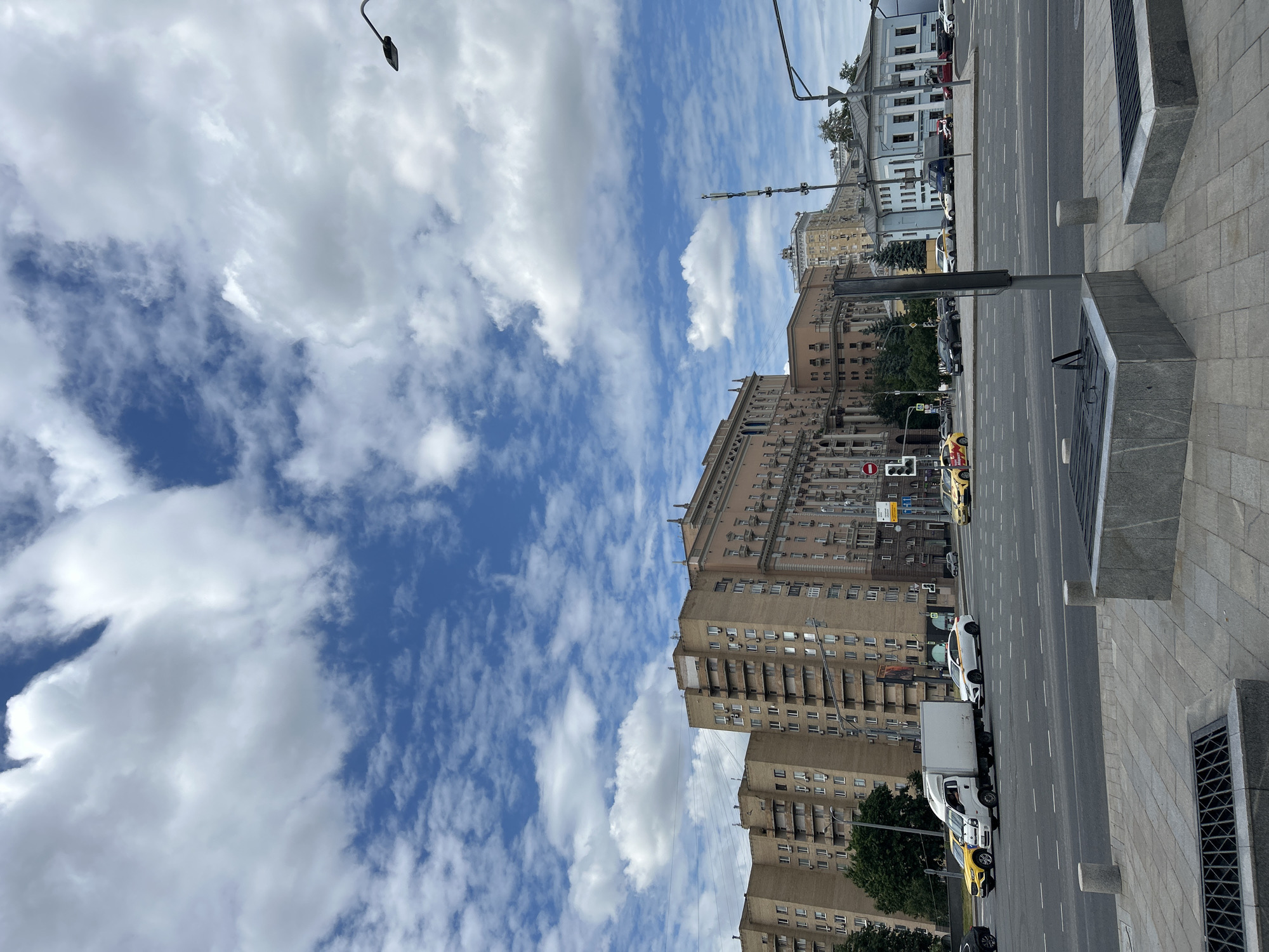 This image depicts a view of the Stalin-era skyscraper, the Ministry of Foreign Affairs building, in Moscow, Russia. The iconic building, characterized by its distinctive architecture and ornate details, dominates the background. In the foreground, a busy street scene unfolds with vehicles, including cars and a white truck, navigating an intersection under a sky filled with dramatic clouds. The street is paved with asphalt and marked with lane lines. The presence of streetlights and traffic signals indicates a functioning urban environment. The overall atmosphere is one of a bustling cityscape under a dynamic sky.