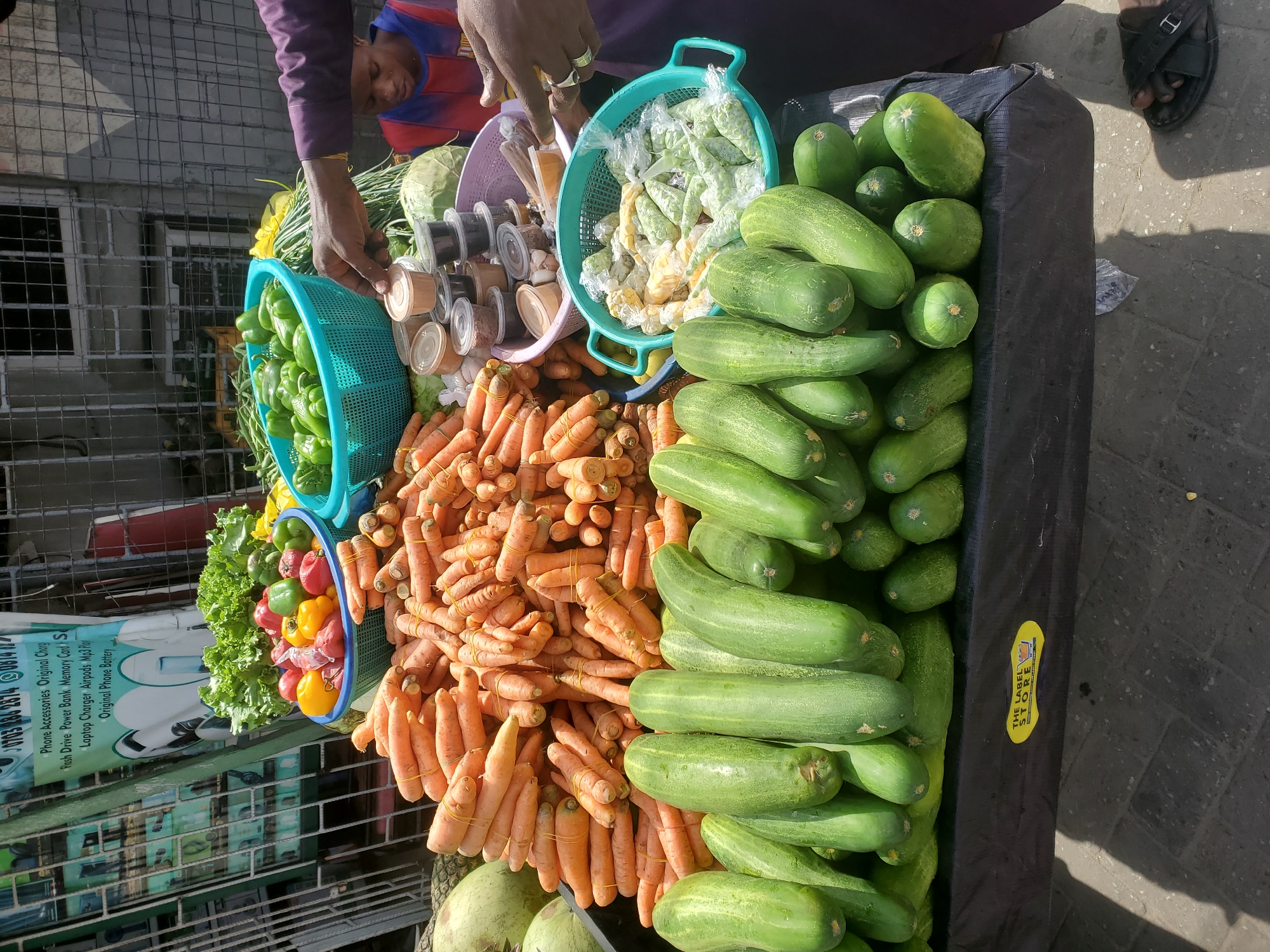This image captures a vibrant street market stall in Agani, Nigeria, bustling with fresh produce under bright daylight. A dark-skinned vendor, wearing a purple long-sleeved shirt and rings on their fingers, is actively engaged in arranging or attending to the goods. Another individual, possibly a child in a striped shirt, is partially visible in the background.
The stall prominently displays an abundance of fresh vegetables. Large piles of bright green cucumbers and orange carrots dominate the foreground, with the carrots neatly bundled together with rubber bands. Various other vegetables are arranged in blue and purple plastic baskets, including green, red, and yellow bell peppers, leafy green lettuce, and a head of cabbage. Another basket holds small plastic bags filled with what appear to be green beans or peas, and possibly shelled corn or similar grains. Adjacent to the vegetables, several small, clear plastic containers filled with different spices or powders are neatly arranged.
The scene is illuminated by strong natural light, casting distinct shadows on the paved ground, indicative of daytime. In the background, a metal fence and part of a building are visible, suggesting an urban market environment. A dark-colored fabric covers the display stand where the produce is laid out.
Visible text on a banner in the background advertises "Phone Accessories," listing various items such as "Original Charg," "Flash Drive," "Power Bank," "Memory Card," "Laptop Charger," "Airpods," "Mp3 Play," and "Original Phone Battery." A yellow sticker adhered to the display stand proudly reads "THE LABEL STORE."