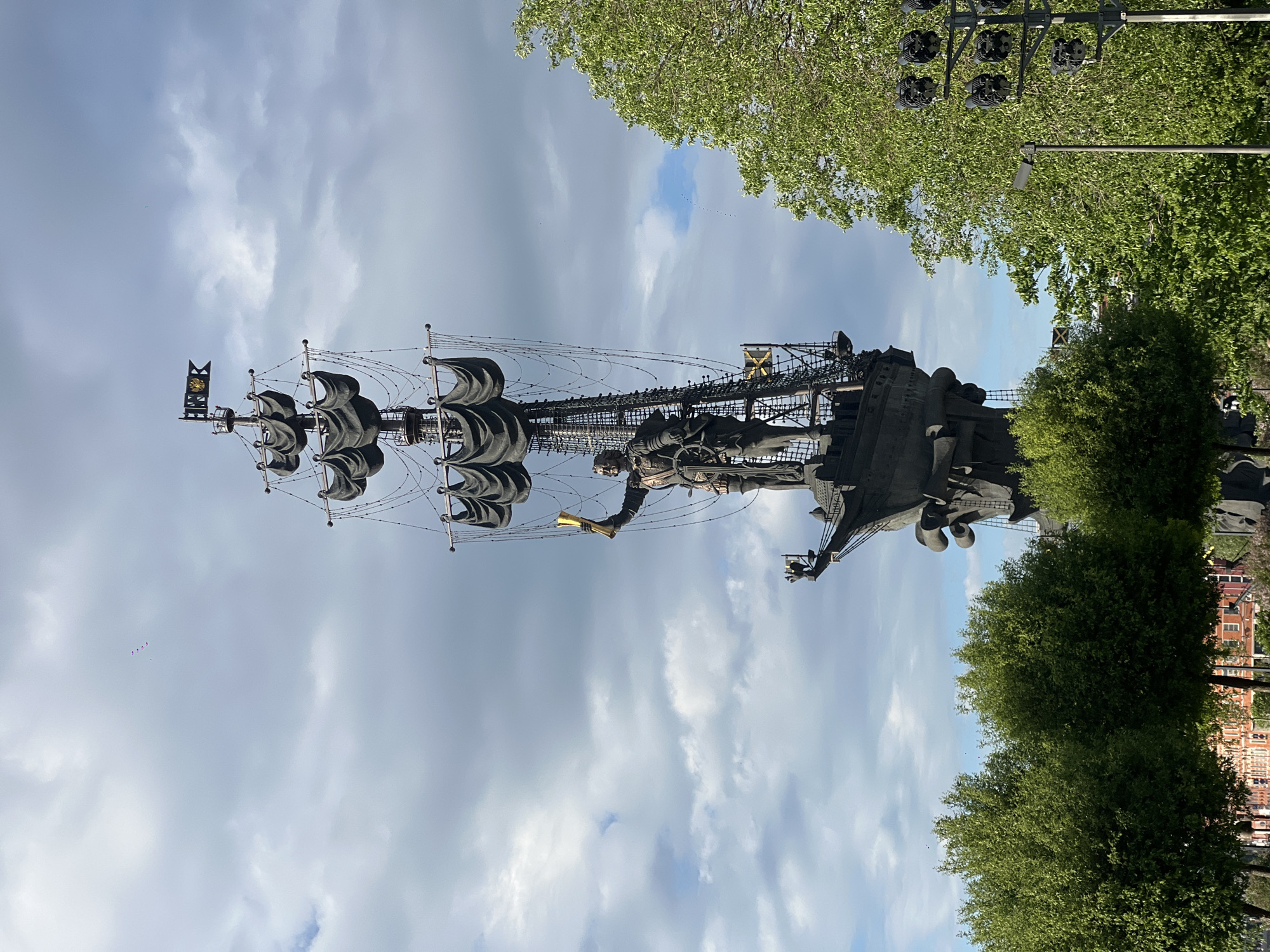 A tall monument of Peter the Great standing on a ship is depicted in the image. The monument is situated in the middle of Moscow, Russia, on the Moskva River. The sculpture is black and features Peter the Great holding a golden scroll. The ship features masts and black sails. The sky is cloudy. Green trees are on the right side of the picture. Buildings can be seen in the far distance.