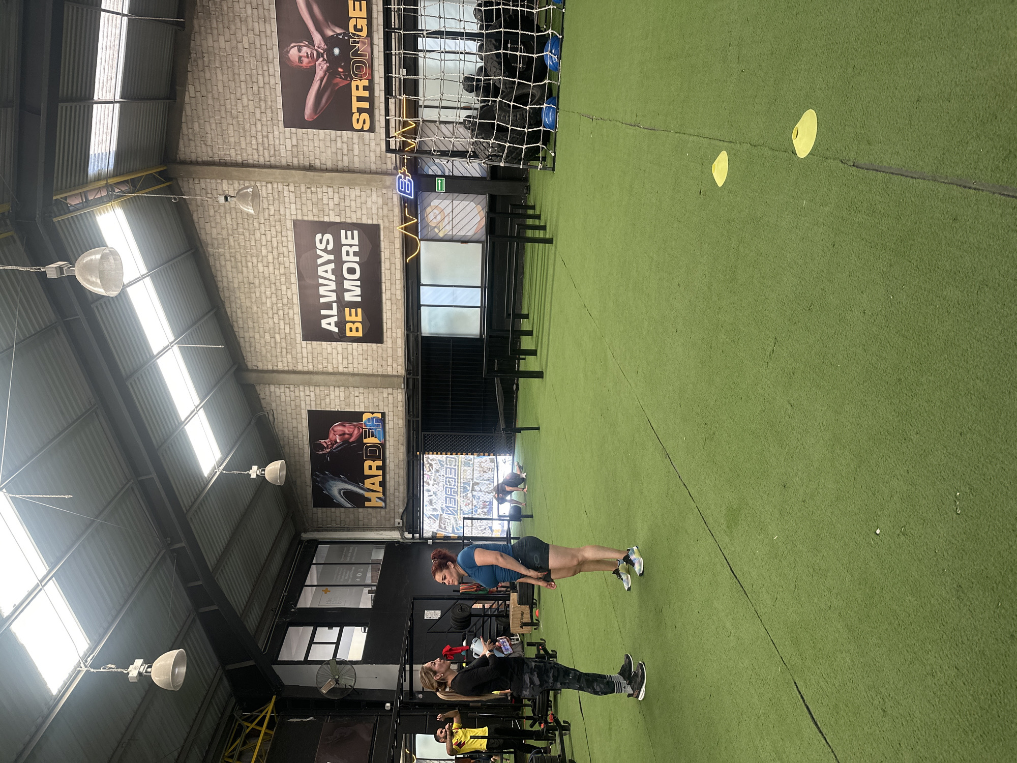 The photo was taken inside a gym in Puebla, Mexico. The gym features a green turf floor, white brick walls with motivational posters ("Stronger," "Always Be More," "Harder"), and a metal ceiling with hanging lights. In the foreground, two women are standing on the turf, one in a plank position, and the other with a water bottle. In the background are gym equipment like weights, a net filled with exercise balls, and more people. Two yellow cones are lying on the turf.