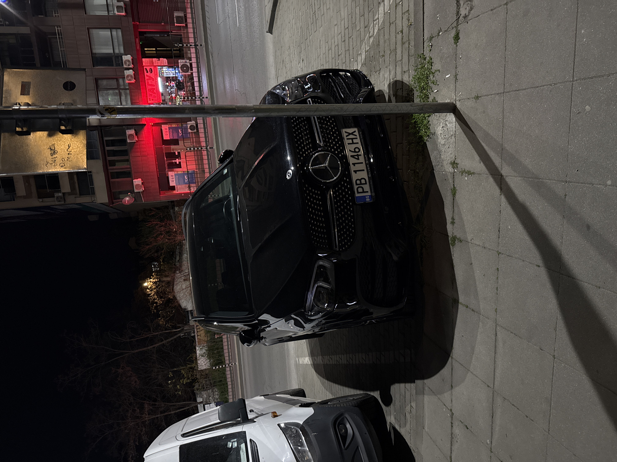 The image depicts a nighttime street scene in Plovdiv, Bulgaria. A black Mercedes-Benz car is centrally parked on a paved sidewalk, with a metal pole partially obscuring its front. The car's grille features the prominent Mercedes-Benz logo, and its Bulgarian license plate, "PB 1146 HX," is clearly visible, with the frame indicating "Mercedes-Benz Silver Star www.silverstar.bg." To the left of the Mercedes, a white commercial van is also parked, its front end and headlights visible.
In the background, multi-story buildings are illuminated by artificial lights, with one building emanating a reddish glow from a storefront. A red neon sign with Cyrillic text, including "АТУ" (ATU), is visible on this building. Below it, several square panels bear the "psd" logo. Multiple air conditioning units are mounted on the building facades. On a metallic panel, graffiti spells out "Juss & More."
The sidewalk is composed of large paving slabs, with small patches of weeds growing through the cracks. A long, distinct shadow cast by the metal pole stretches across the pavement. Beyond the immediate street scene, bare trees are silhouetted against the dark night sky, with a single distant streetlight providing minimal illumination. The overall scene conveys a quiet, urban environment after dark, with parked vehicles suggesting residential or commercial activity.