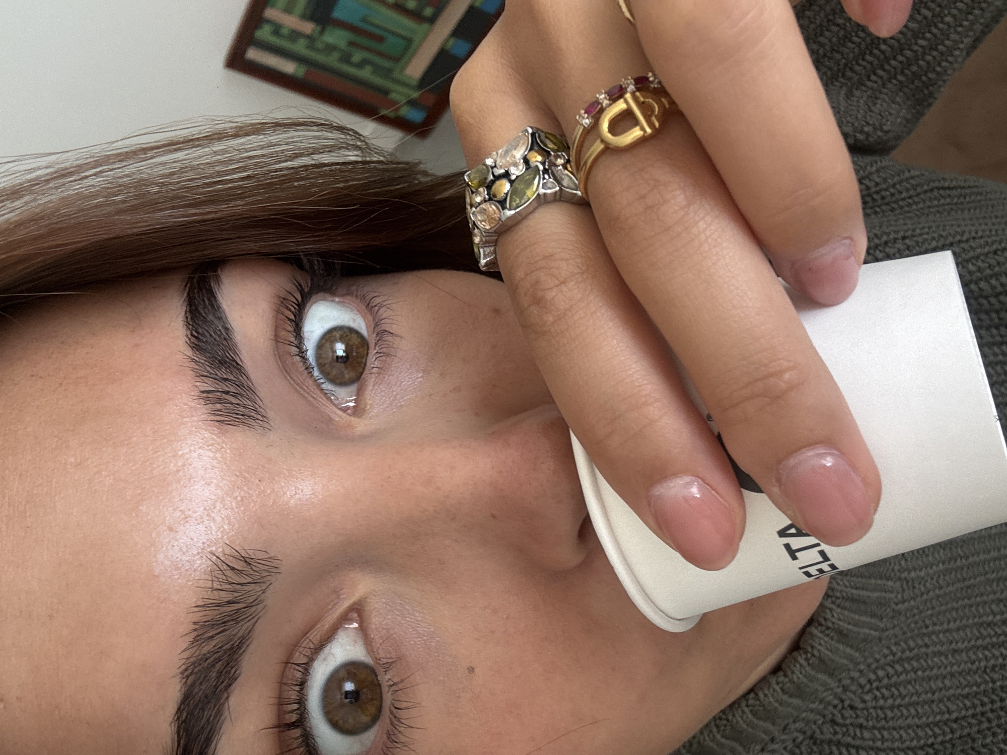 A close-up, tilted image shows a person holding a white paper cup with the letters "ELTA" partially visible. The person has brown eyes and dark eyebrows. They are wearing several rings, including a silver ring with green and clear stones, and a gold ring with small red stones. The person is wearing a gray-green textured sweater. In the blurred background, a decorative wall hanging with geometric patterns is partially visible. The location cannot be determined to be Torres Vedras, Portugal from the visual information provided.